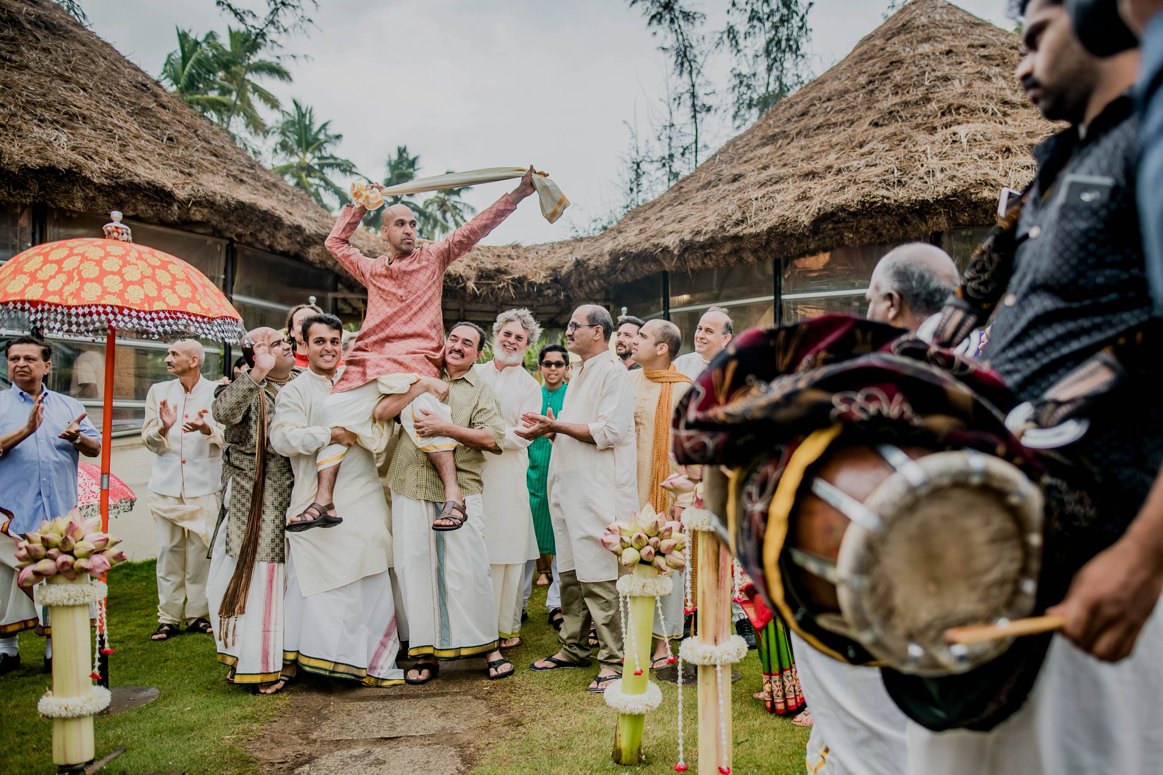 Groom in pink raises arms joyfully during outdoor ceremony with guests, thatched structures, and traditional drum music