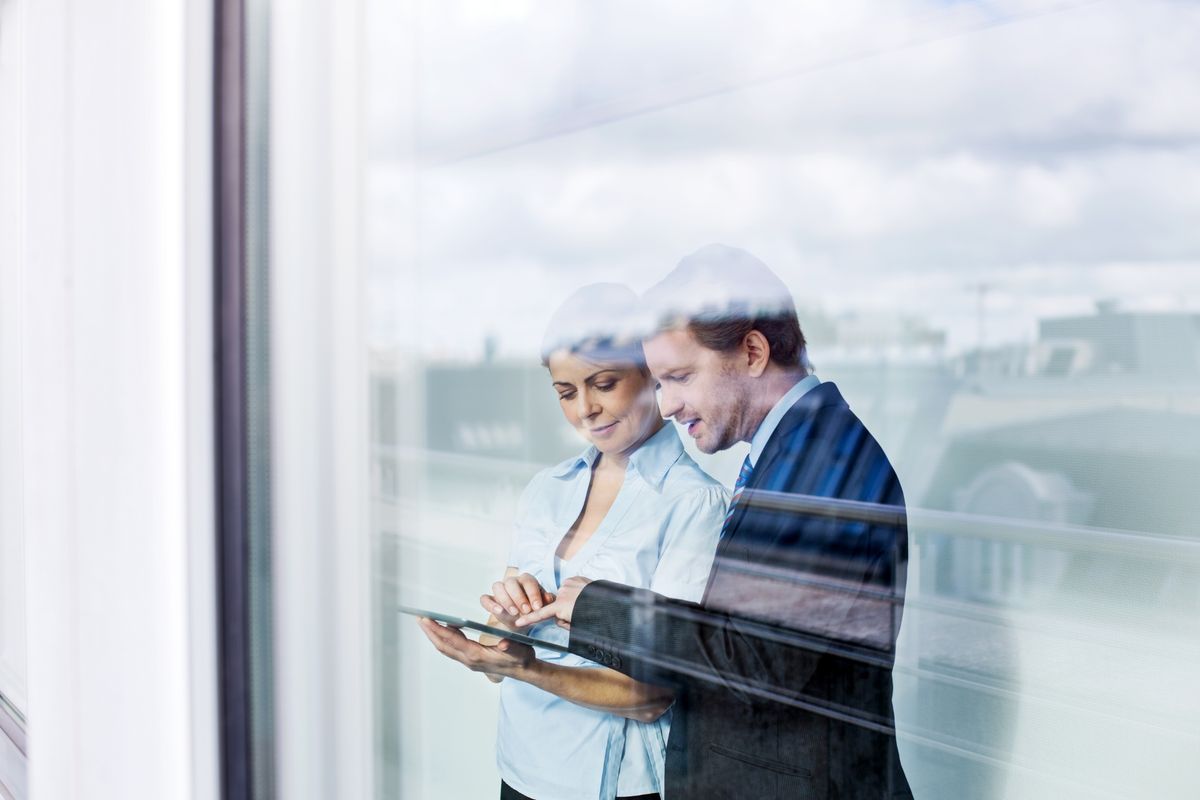 Zwei Menschen im Businessoutfit stehen am Fenster und sind auf ein Tablet fokussiert, was eine der Personen in der Hand hält. Das Foto ist von außen aufgenommen, in der Scheibe spiegelt sich eine Stadtsilhouette. // Two people in business attire stand at a window, focused on a tablet, that one of them is holding. The photo was taken from outside, with a city skyline reflected in the window.