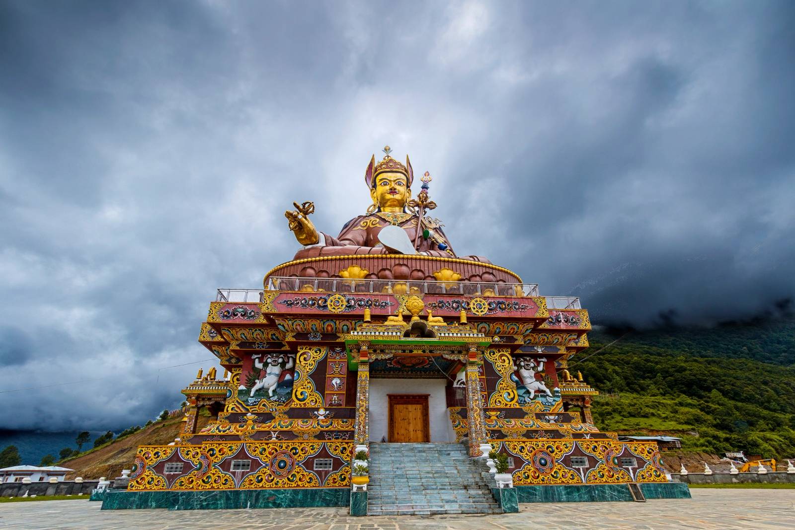 The giant gilded statue of Guru Rinpoche seated on its painted temple base at Takila, under a cloudy sky