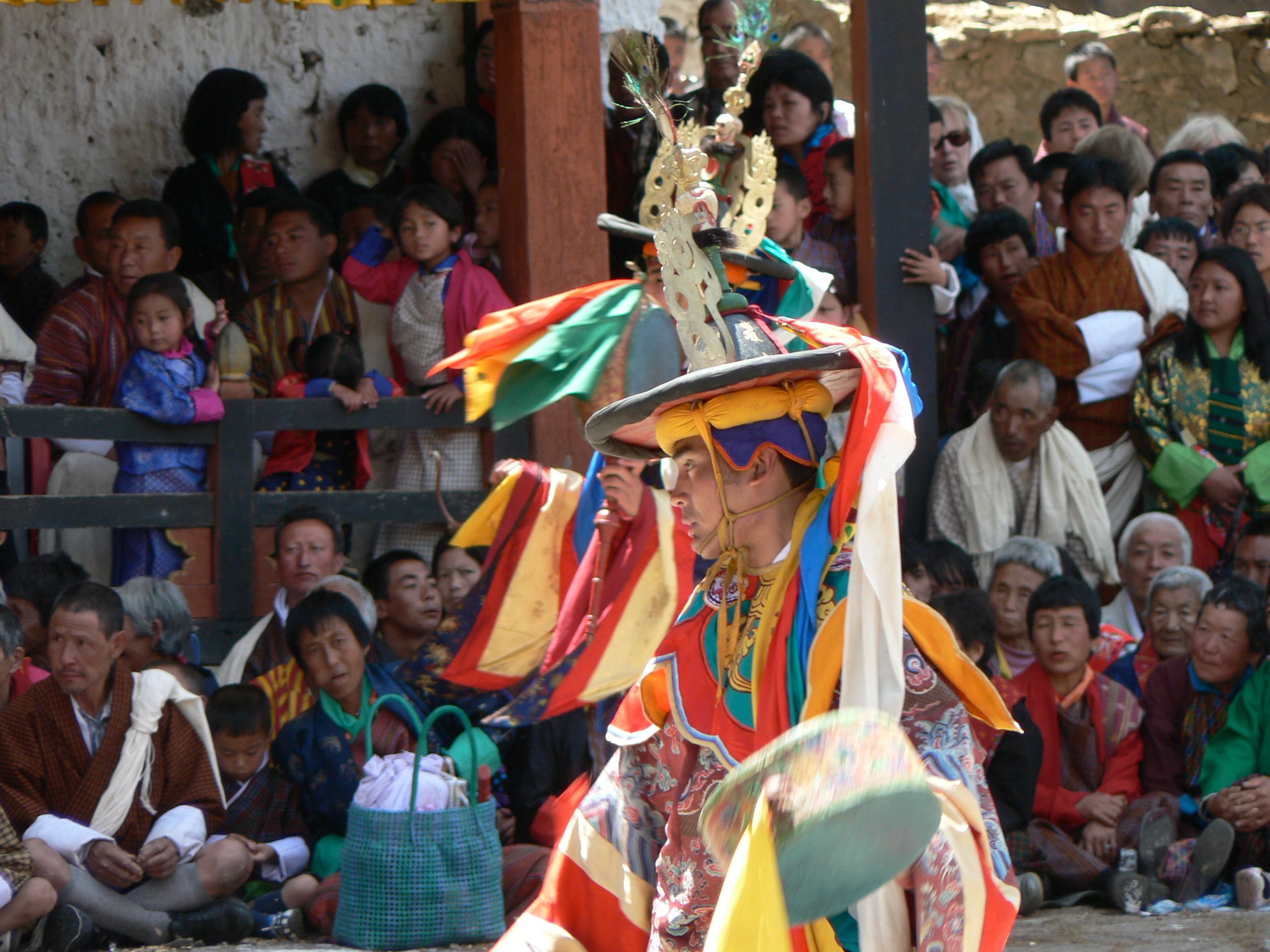 A dancer in a wide-brimmed black hat crowned with gold ornaments and a brocade costume turns mid-dance holding a hand drum, as spectators in gho and kira watch from behind a wooden rail
