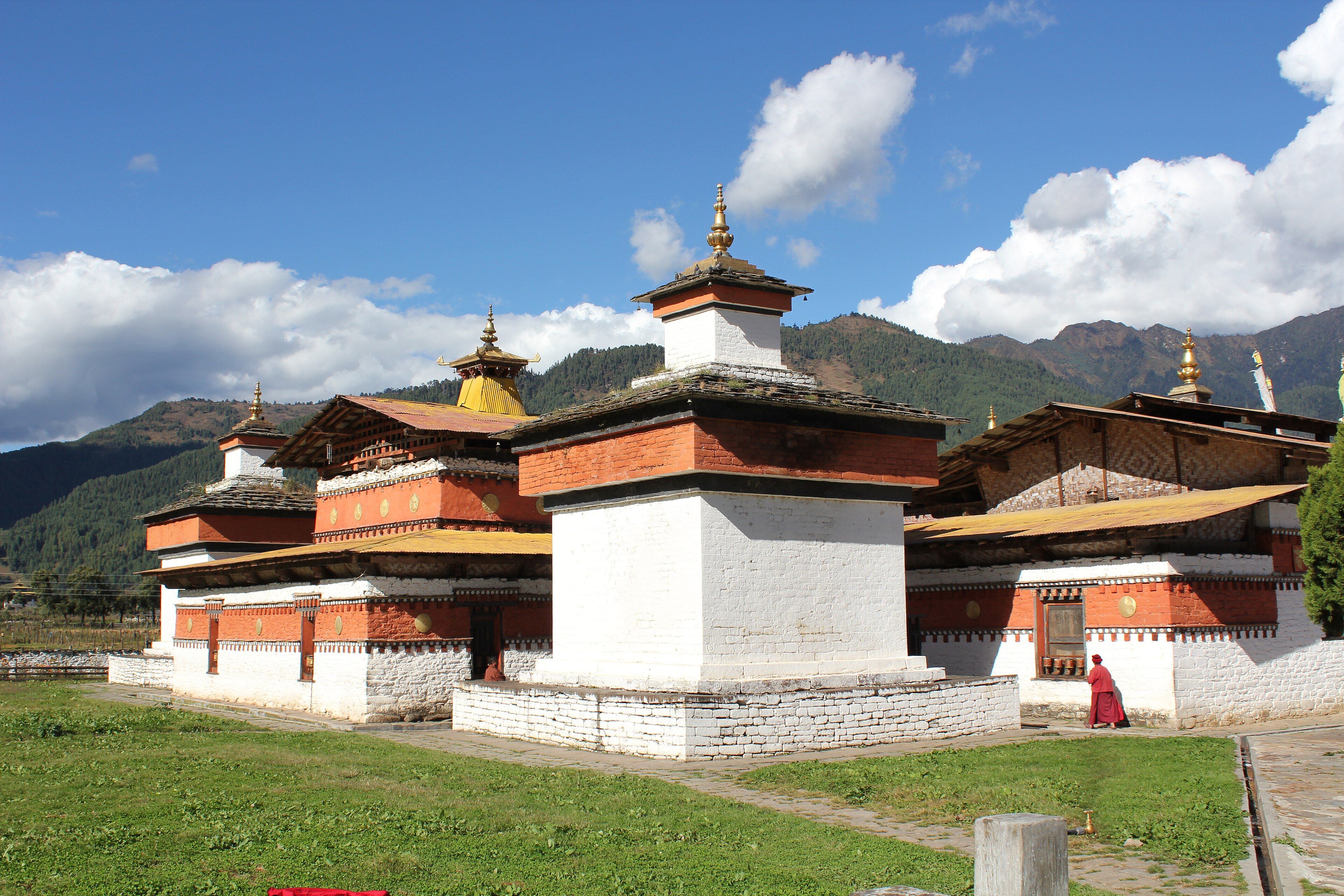 The whitewashed temple buildings of Jambay Lhakhang, with red bands and golden roof finials, on a lawn as a monk in red robes walks past.