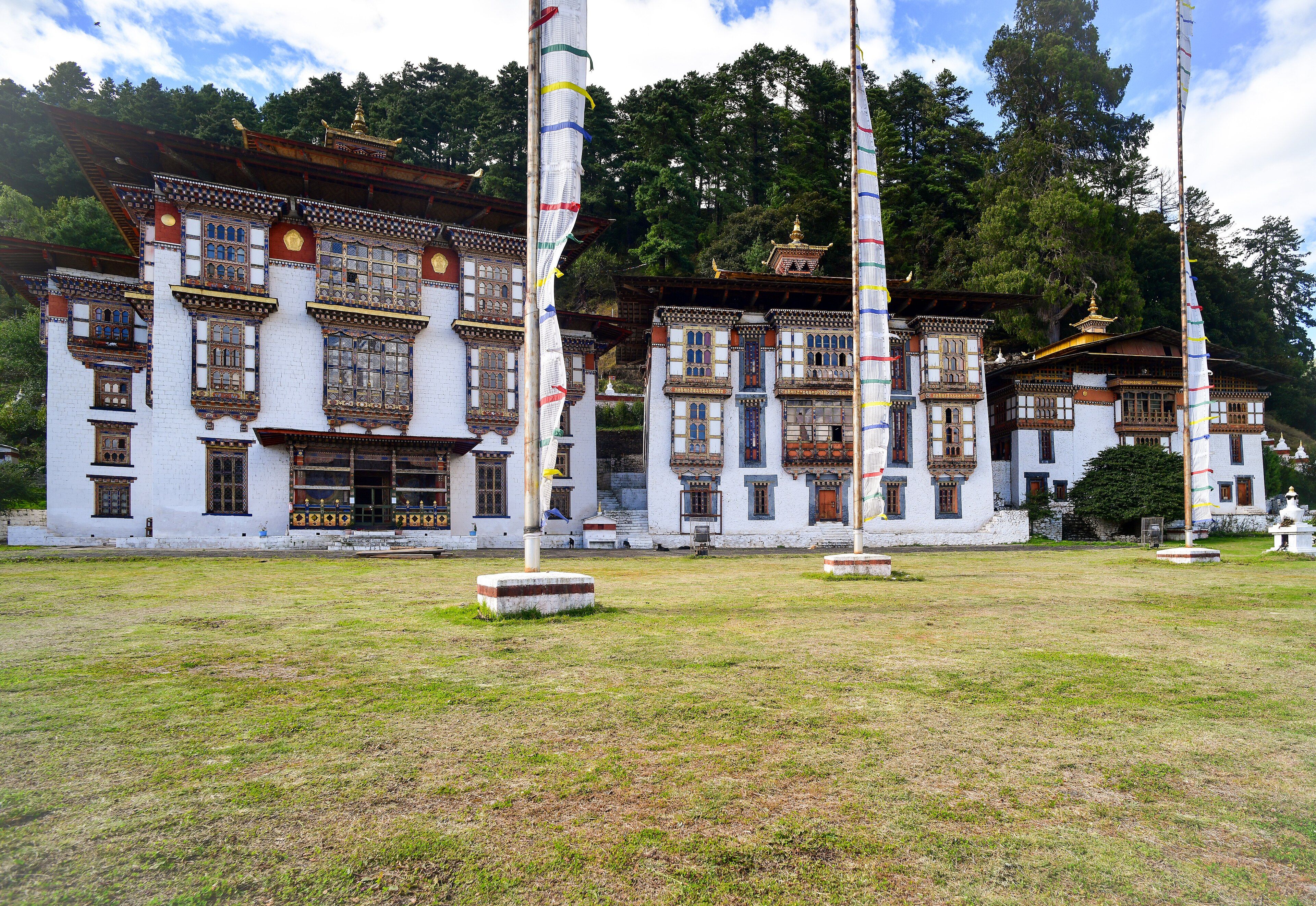The three white temple buildings of Kurjey Lhakhang against a forested slope, with tall prayer-flag poles standing on the grass courtyard in front.