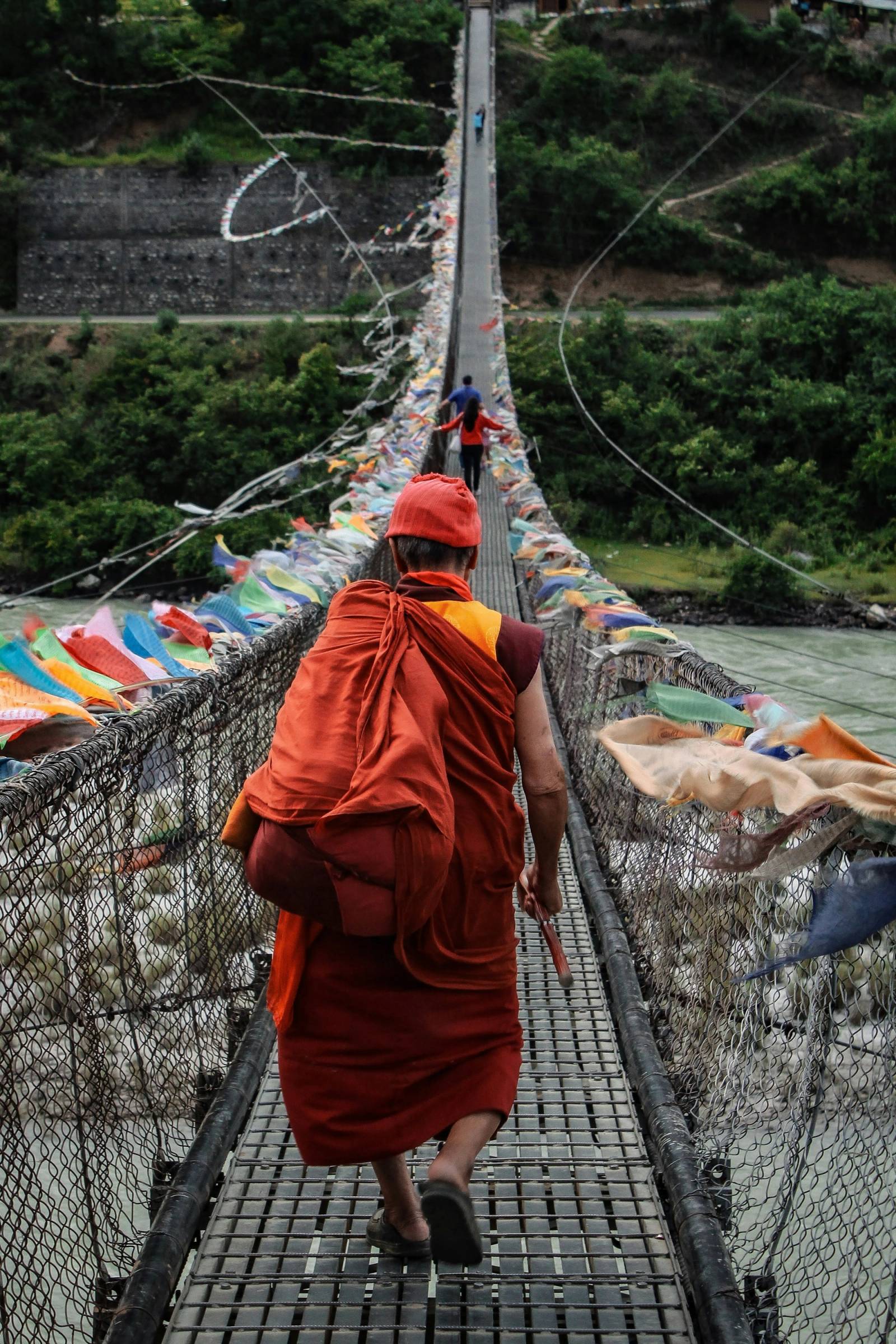 A monk in red robes crosses a long suspension bridge hung with prayer flags