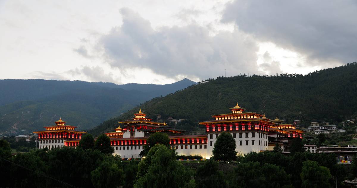 Tashichho Dzong's long whitewashed walls and golden roofs, its windows lit red at dusk, below forested hills and a cloudy sky