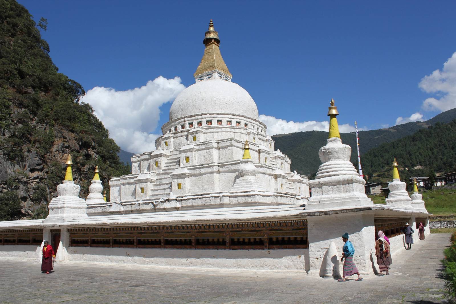 The white dome and gilded spire of Chorten Kora, with pilgrims walking round it under a blue sky