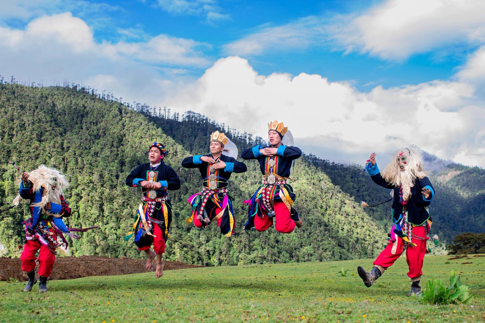 Brokpa dancers in masks, crowns and red trousers leaping on a highland meadow below forested hills
