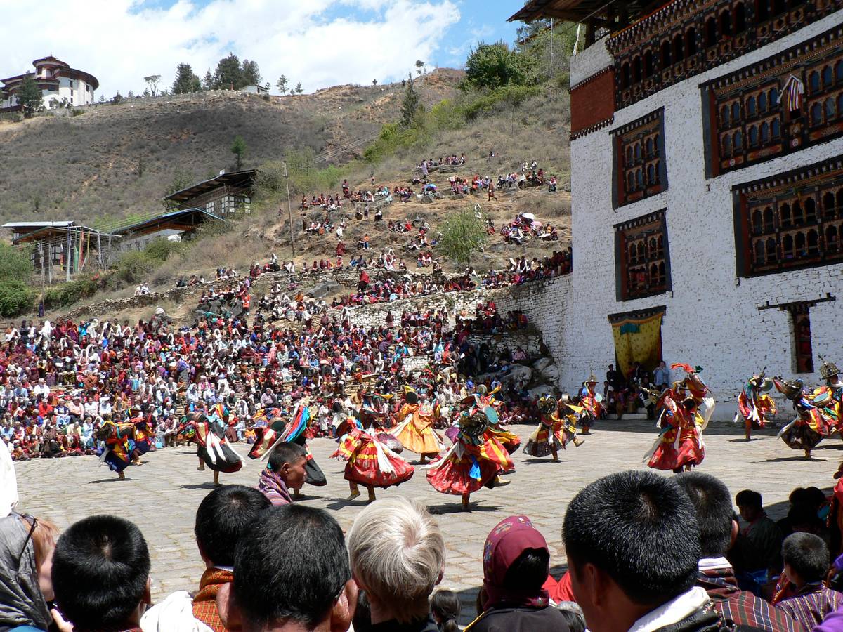 Dancers in black hats and red and gold costumes whirl across a stone-paved ground beside the white walls of a dzong, watched by a crowd packed onto stone terraces and the dry hillside above