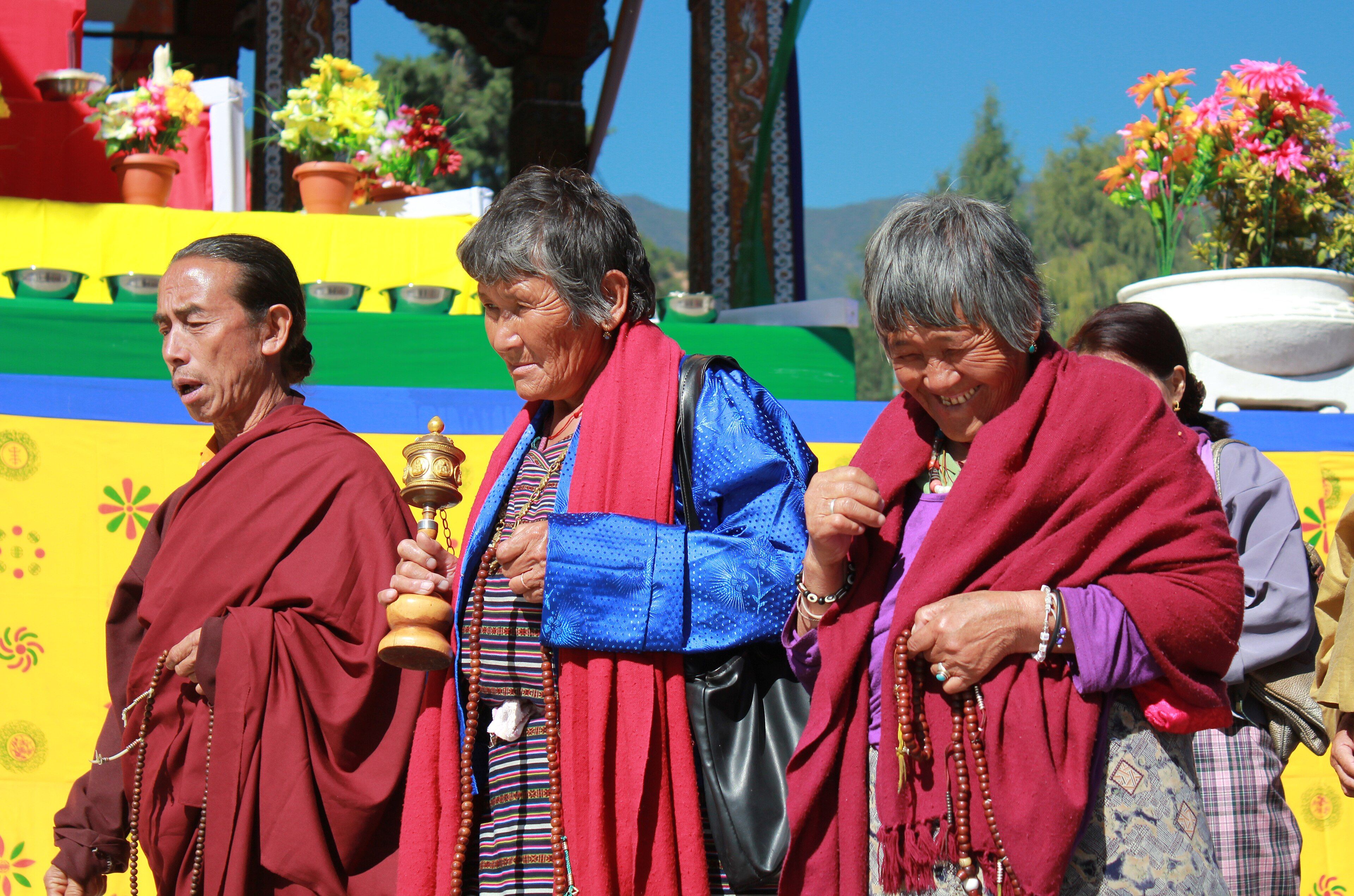 Elderly women in red shawls and a man in maroon robes walk past flower offerings at the Memorial Chorten, one turning a hand-held prayer wheel, all holding prayer beads