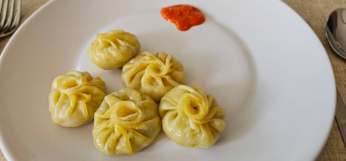 Five steamed momos with pleated tops on a white plate, with a spoonful of orange-red chilli sauce beside them.