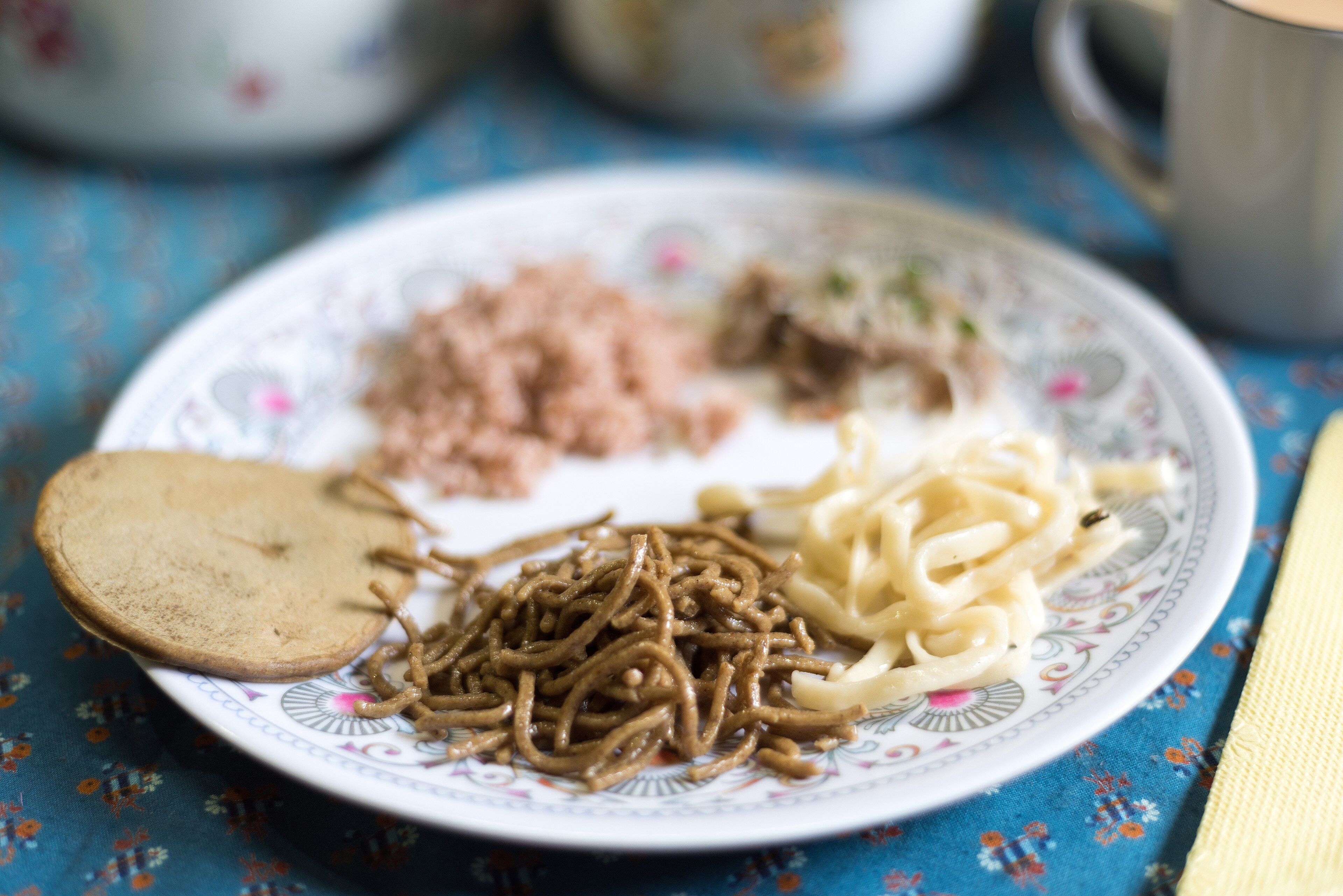 A plate with a round buckwheat pancake, a pile of brown buckwheat noodles, white noodles and pink-red rice, next to a mug.