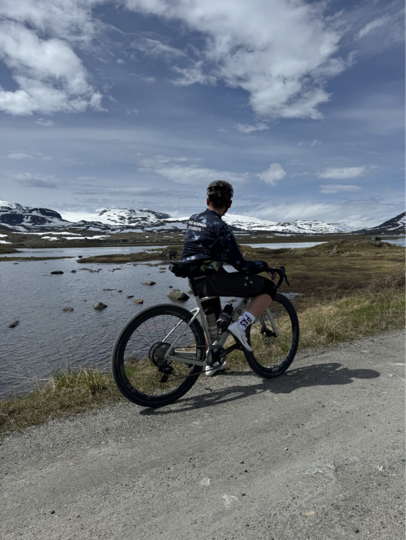 Eirik sykler på en grusvei ved et vann, med snødekte fjell og en høy himmel i bakgrunnen.