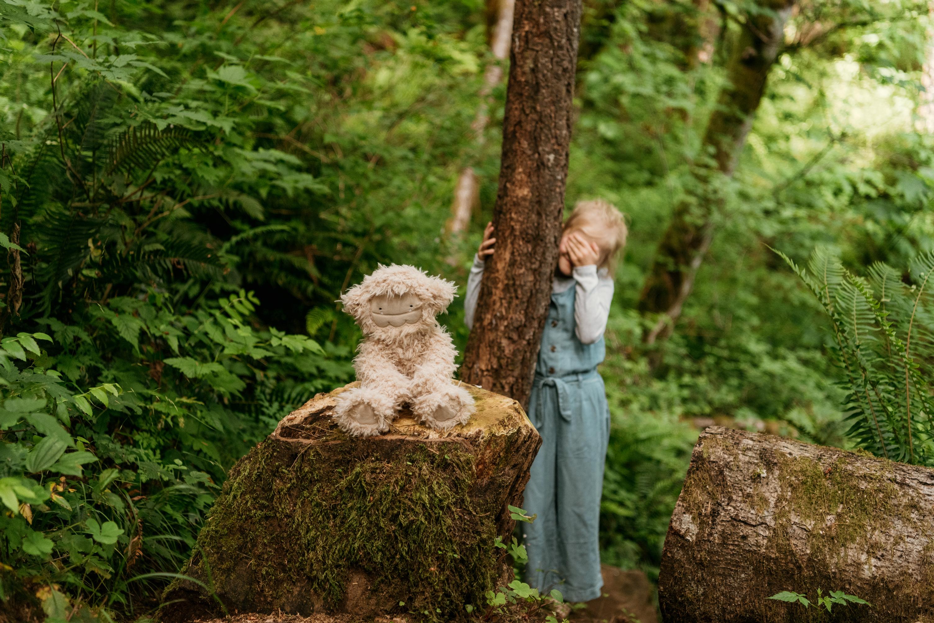 Slumberkins stuffed animal sitting on a stump with a child playing peek-a-boo in the background behind a tree