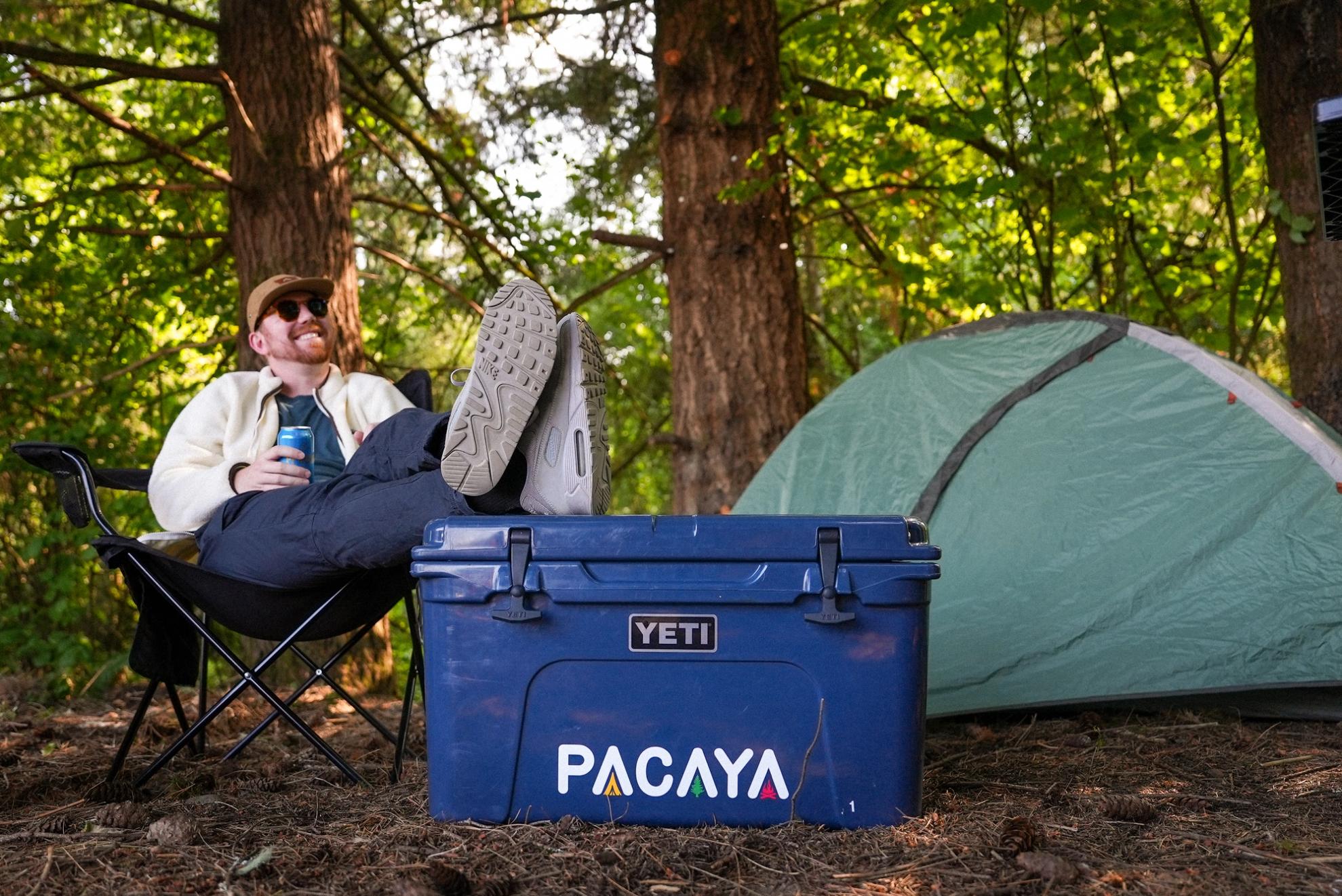 Man sitting with his feet up on a Pacaya cooler, next to a tent, in the woods