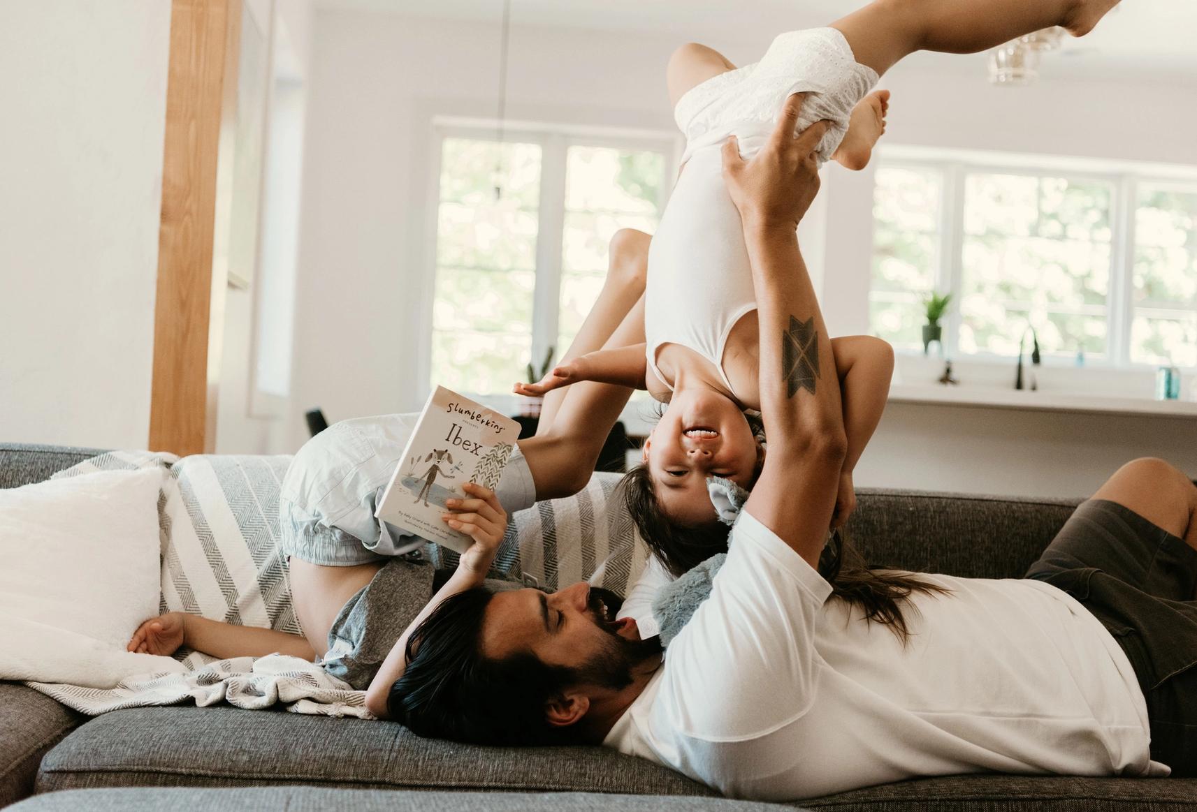 A dad flipping his children on a couch while one of them is holding a Slumberkins book.
