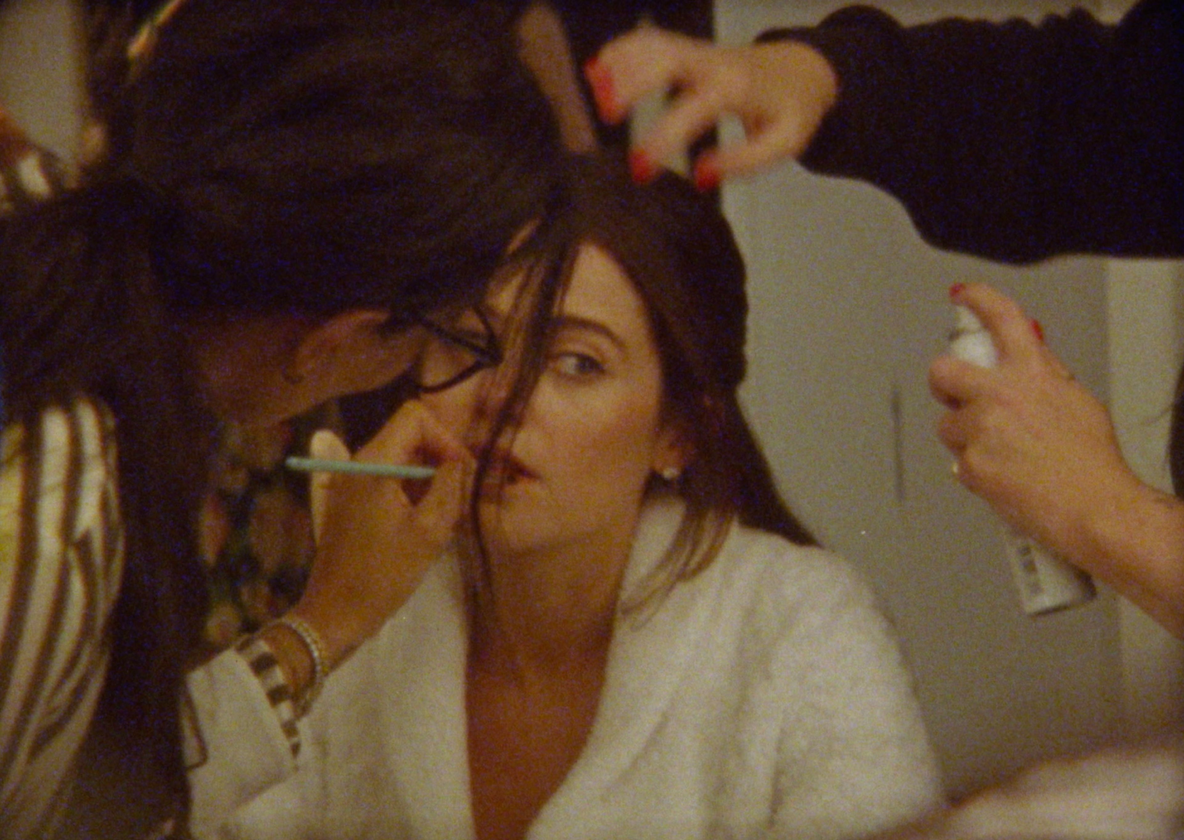 Molly Gordon sits for makeup and hair styling, surrounded by artists mid-touch-up — captured candidly by Sarah Davis.