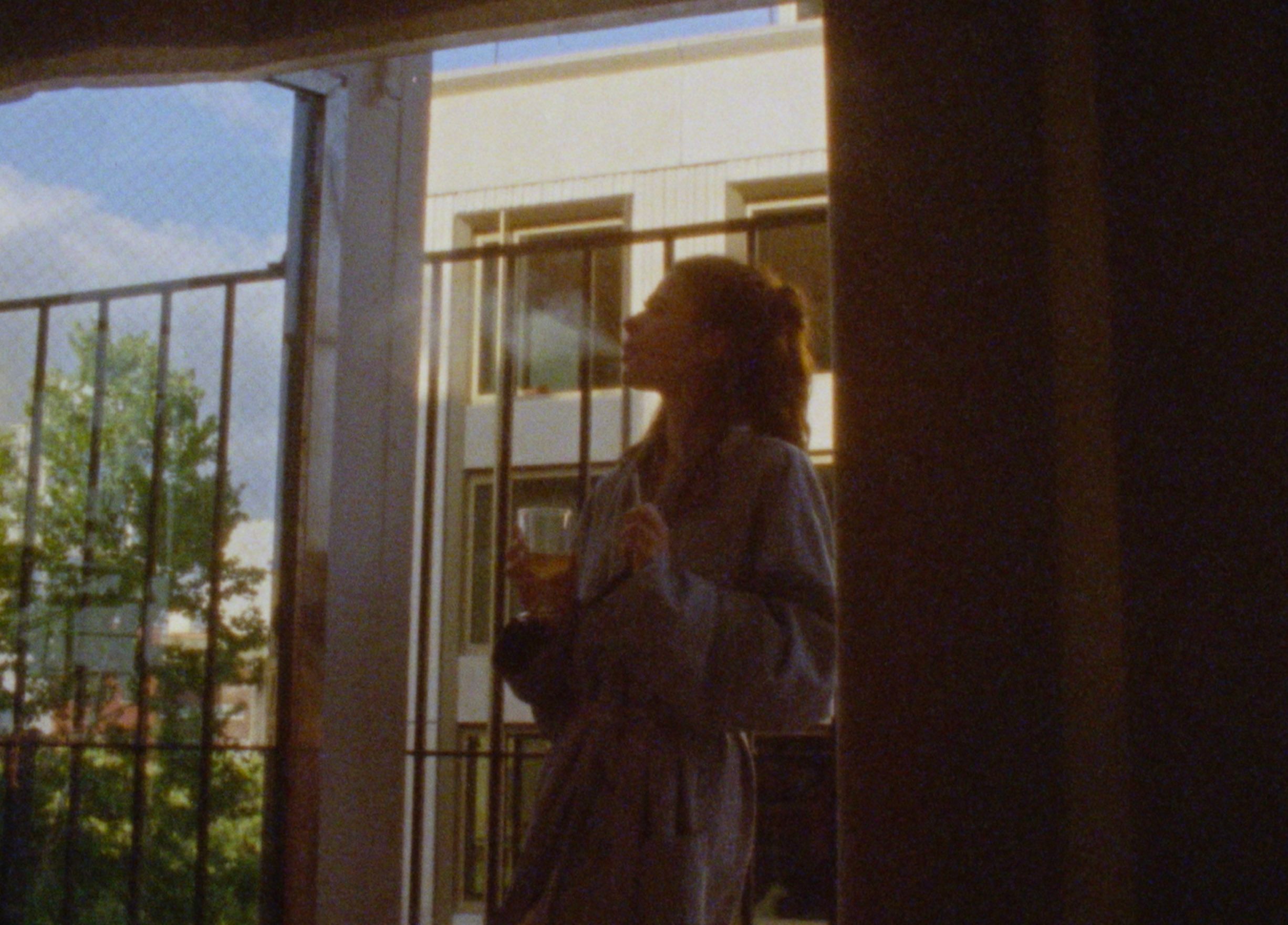 Molly Gordon stands on a balcony in a bathrobe, holding a drink and exhaling softly into the evening light — photographed on film by Sarah Davis.