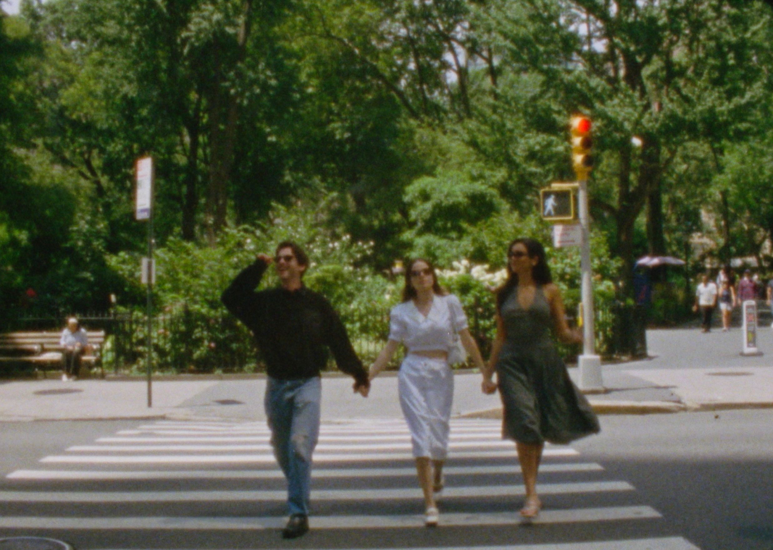 Molly Gordon and two friends walk hand-in-hand through a city crosswalk under summer trees — film still by Sarah Davis.