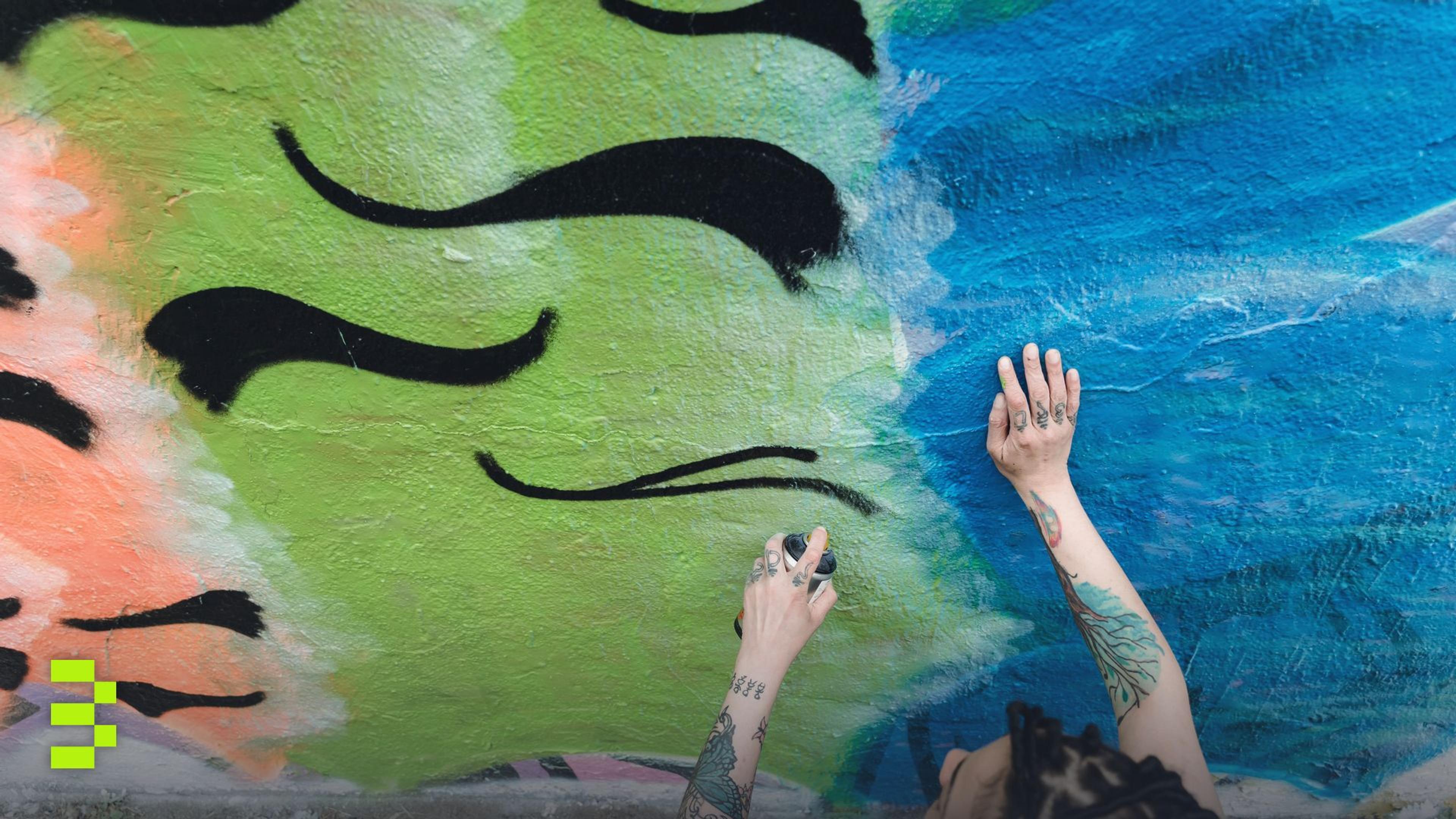A woman with tattoos spray paints a colorful graffiti mural on a concrete wall outside.