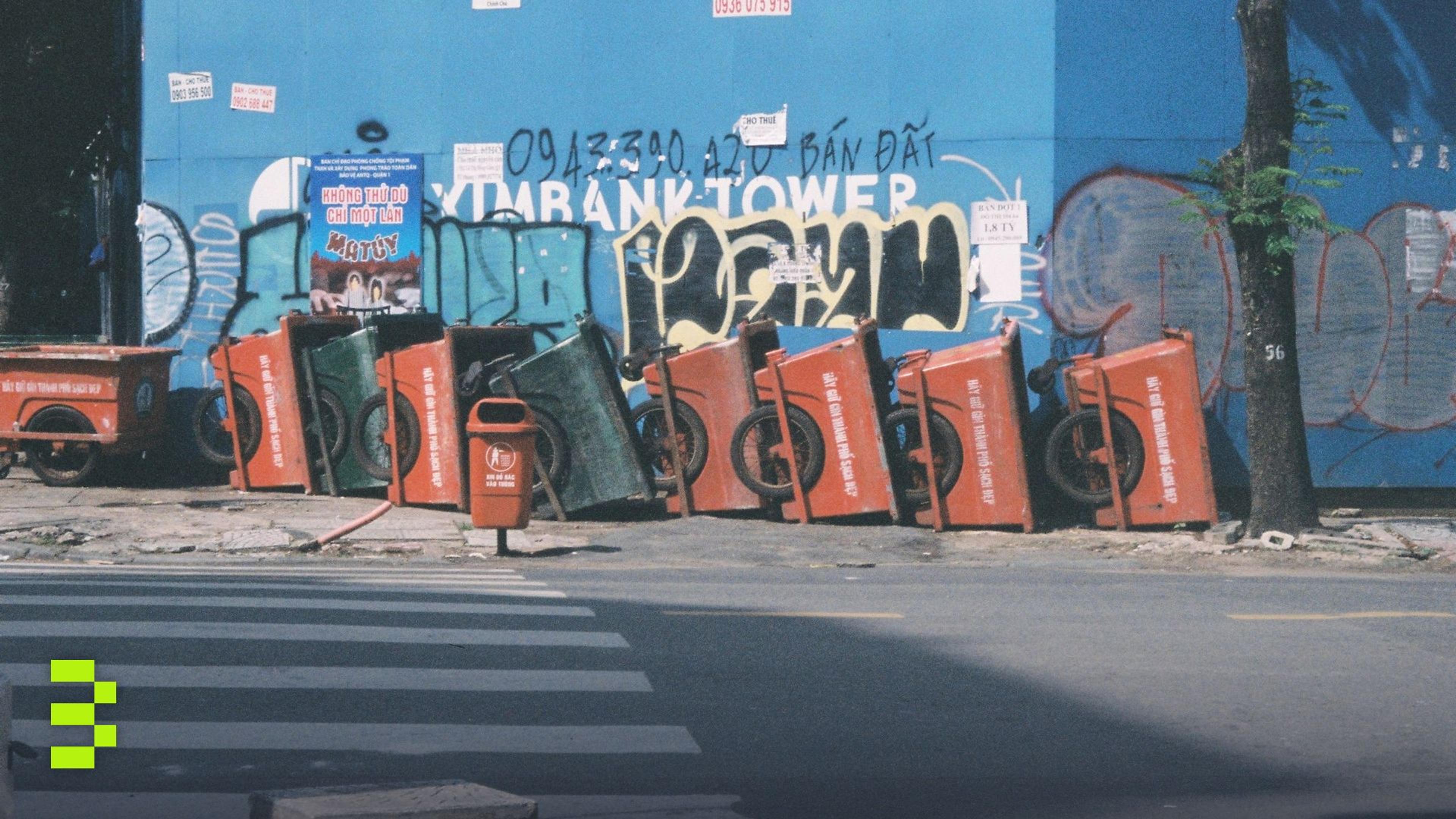Colorful trash bins aligned against a graffiti wall on a Ho Chi Minh City street.