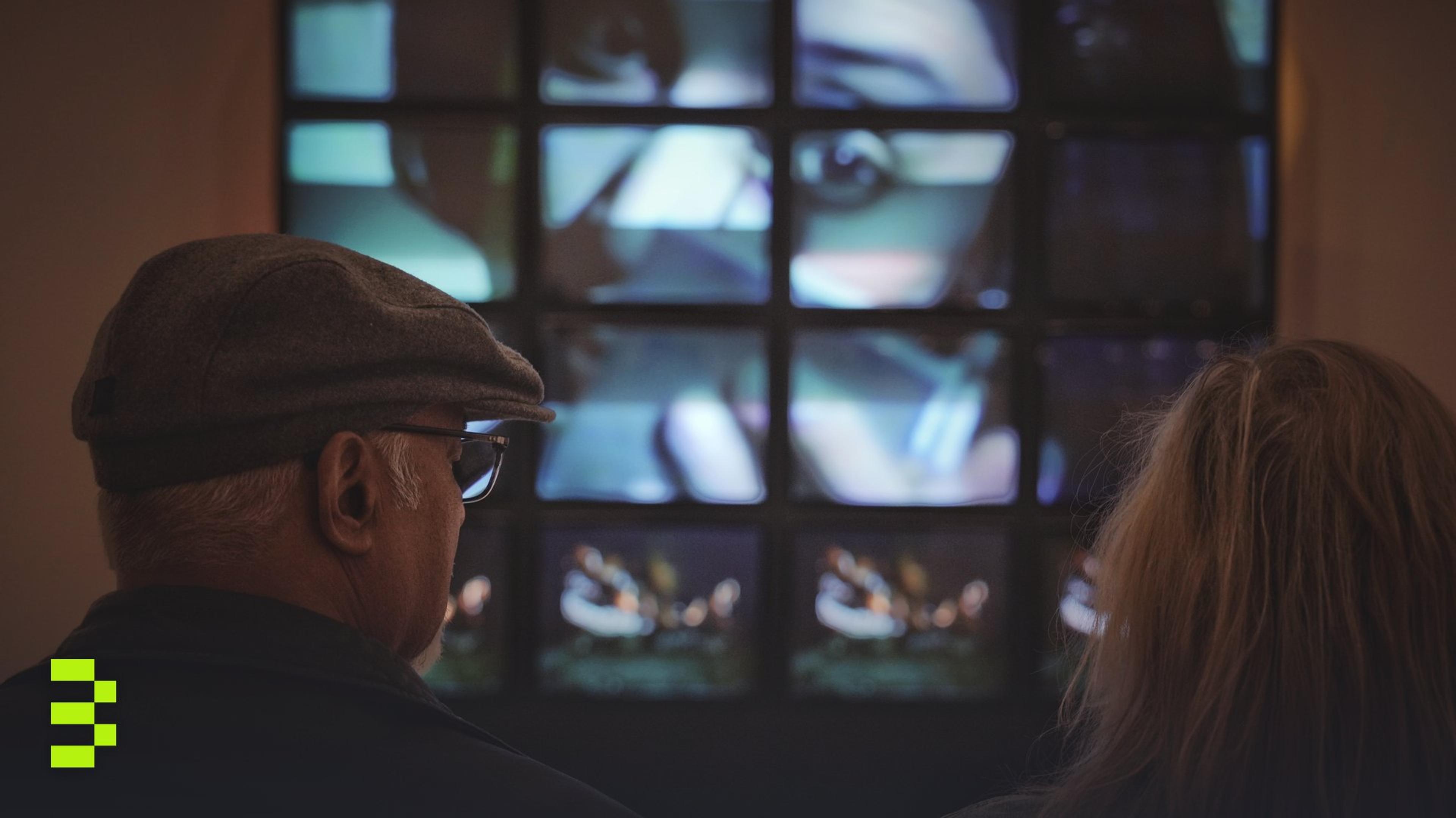 An elderly couple observes a digital art installation on a multi-screen display indoors.