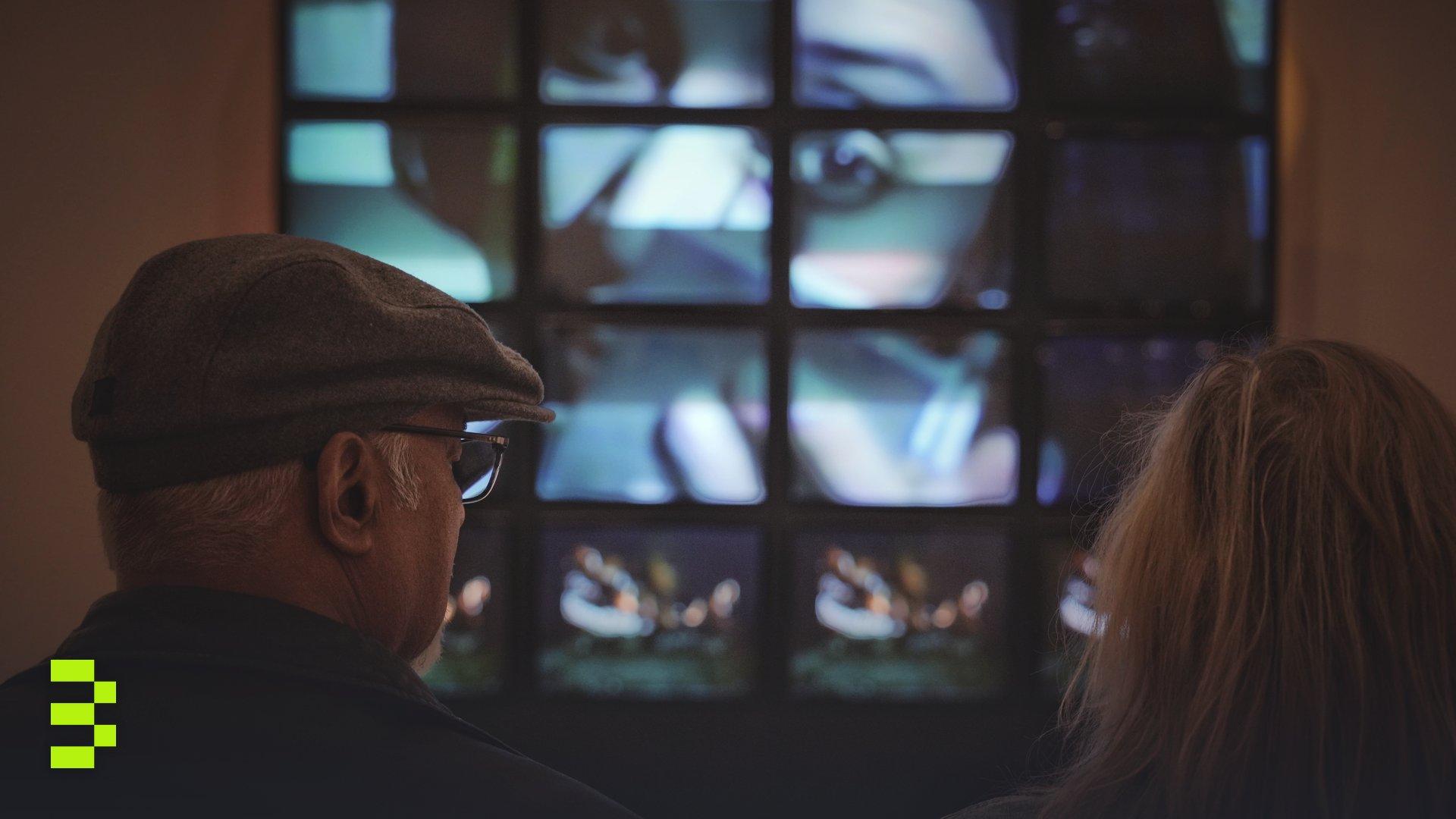 An elderly couple observes a digital art installation on a multi-screen display indoors.