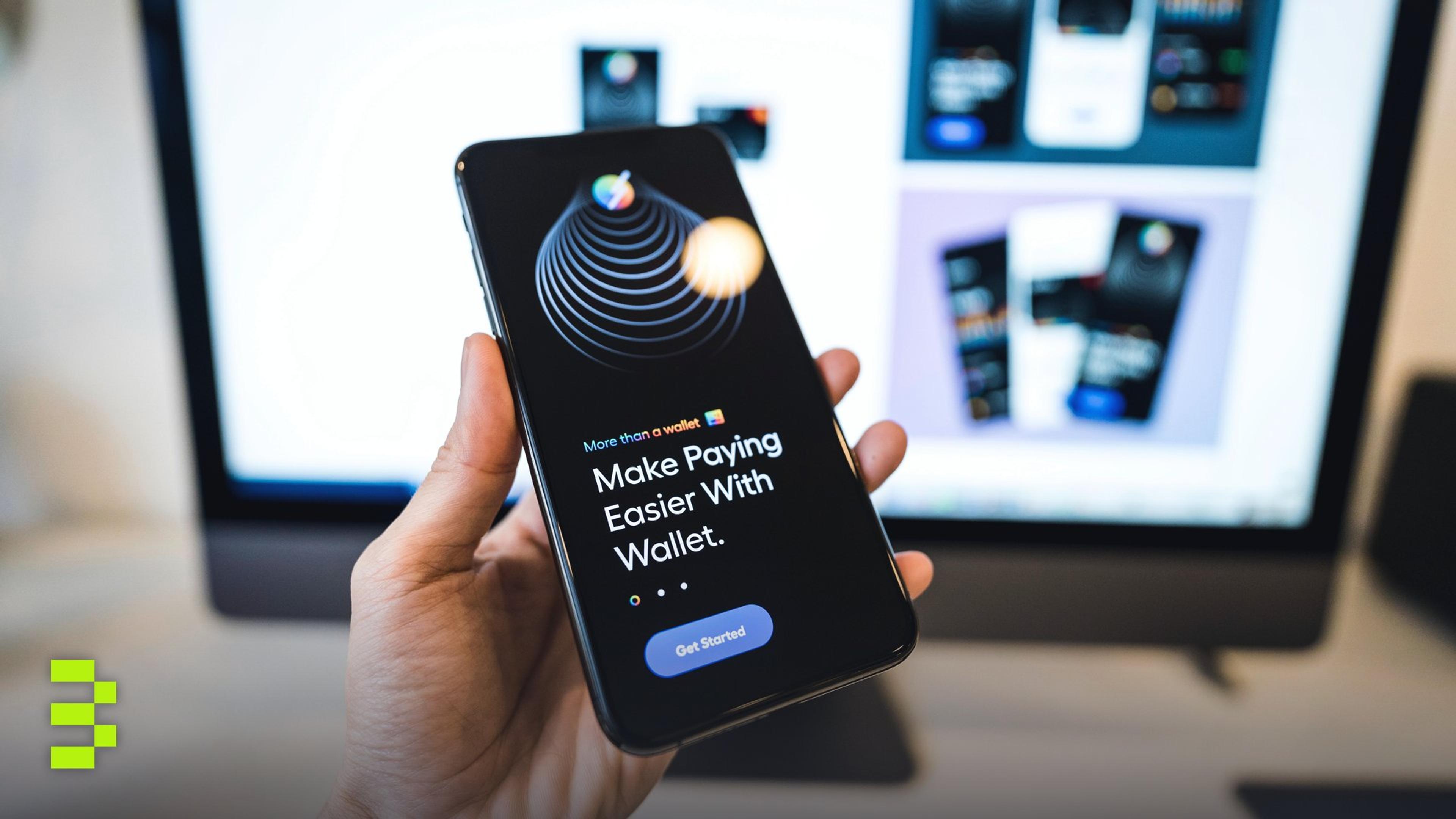Hand holding smartphone displaying digital wallet app interface, blurred monitor in background.