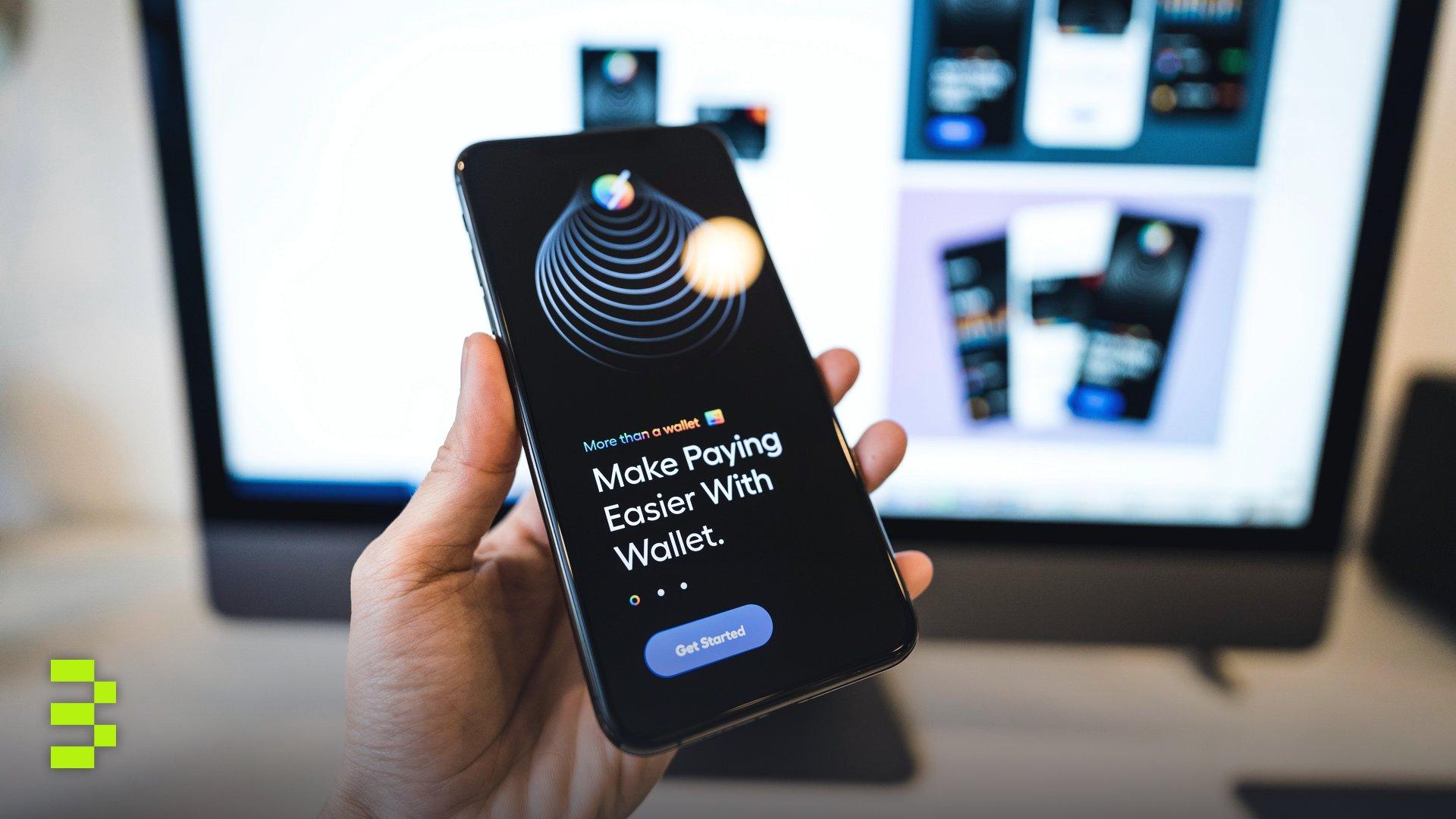Hand holding smartphone displaying digital wallet app interface, blurred monitor in background.
