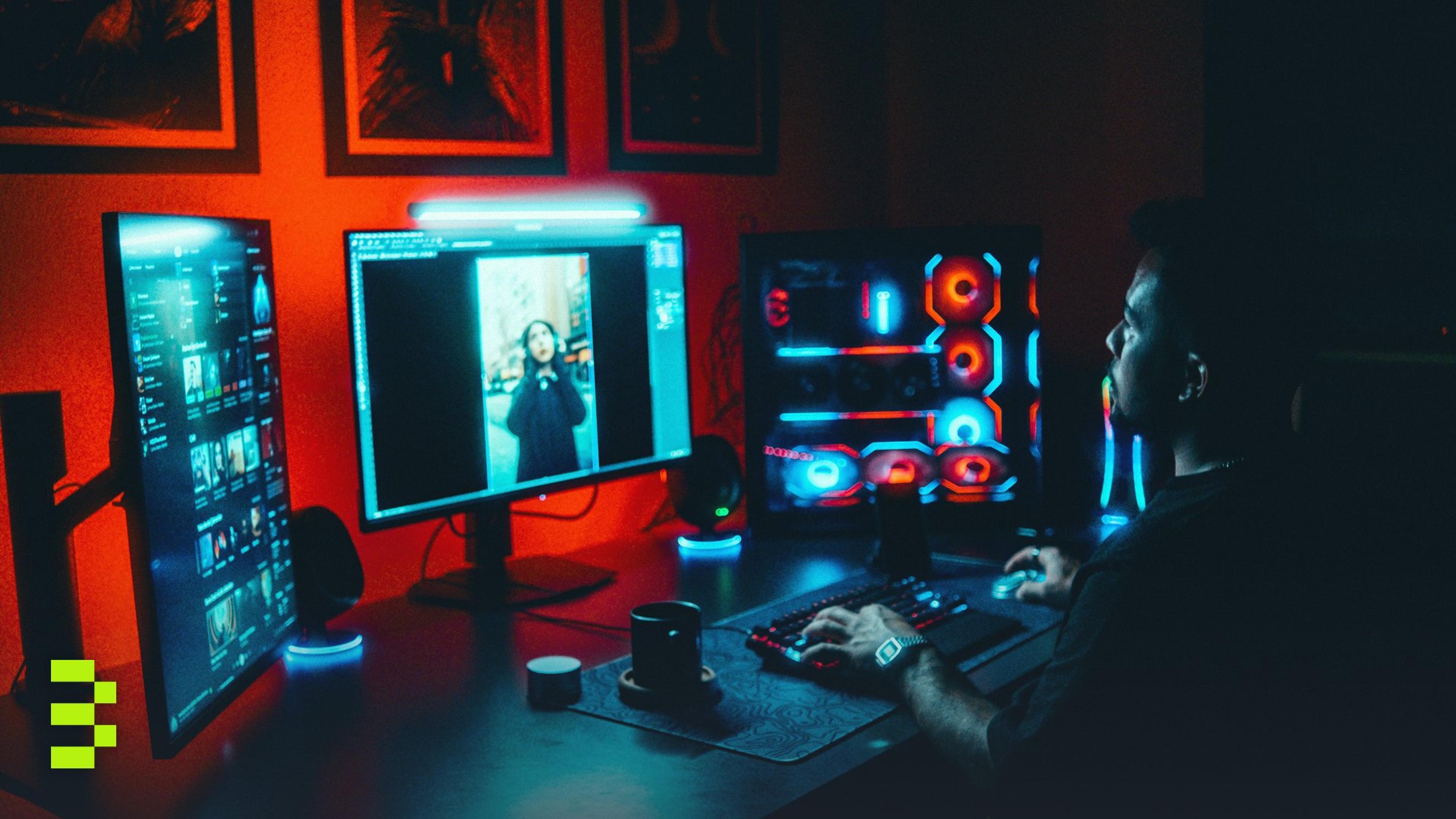A young man working on a multi-screen computer setup in a dark, neon-lit room.
