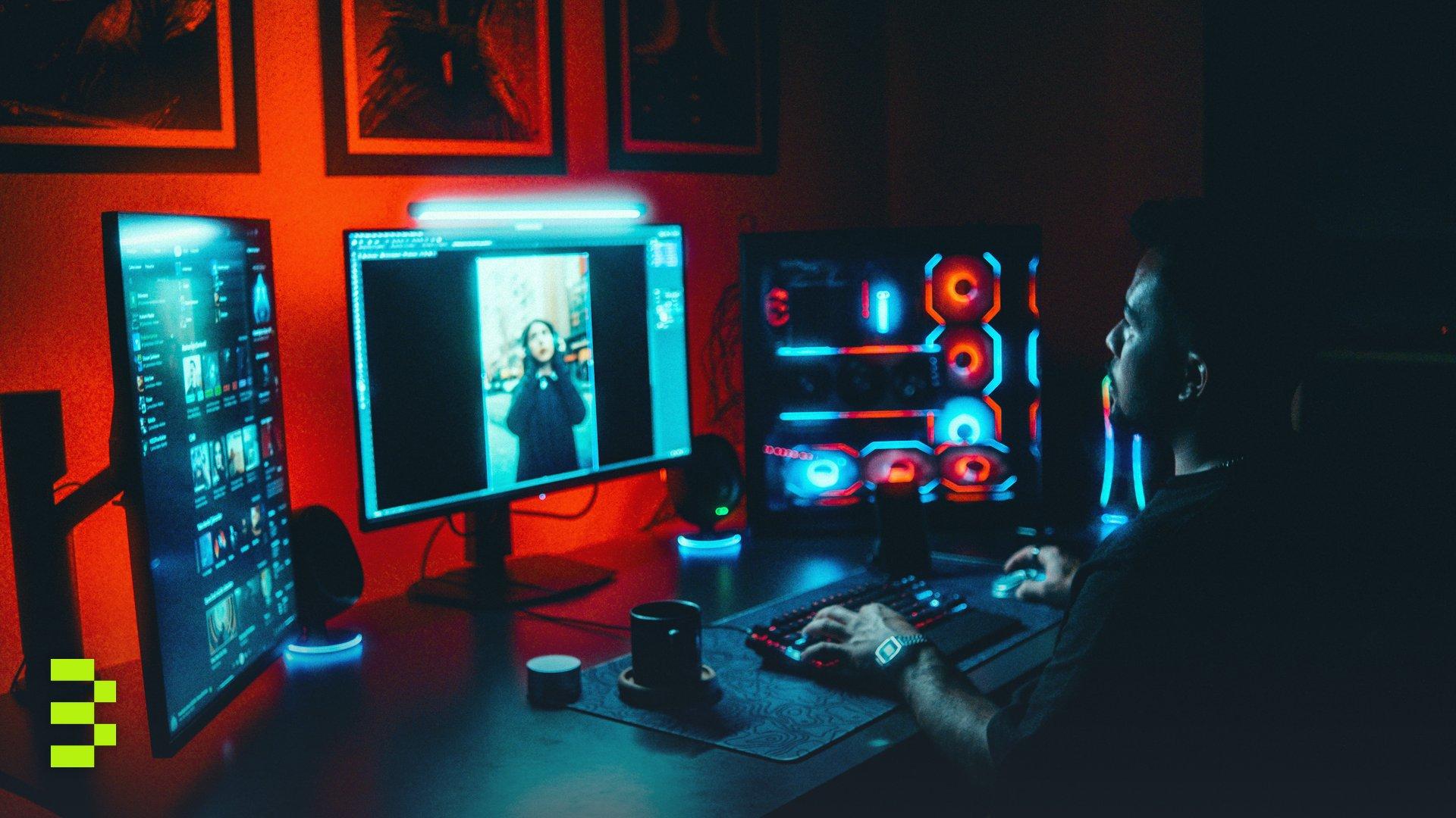 A young man working on a multi-screen computer setup in a dark, neon-lit room.