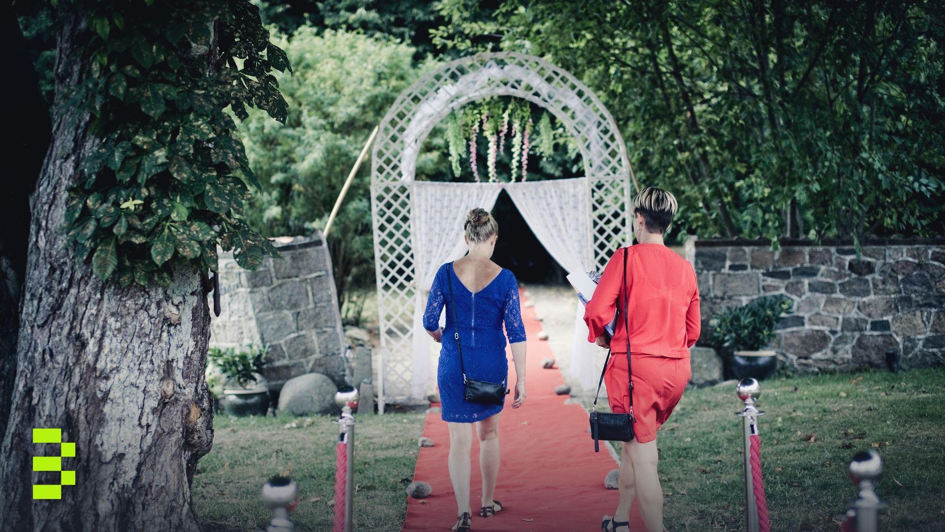 Two women walk on a red carpet through a garden towards a decorative archway.