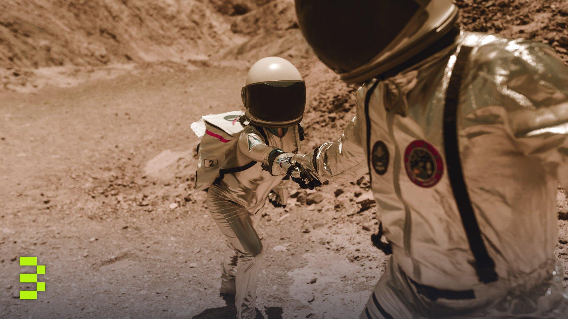 Two astronauts in space suits help each other in a desert-like terrain.
