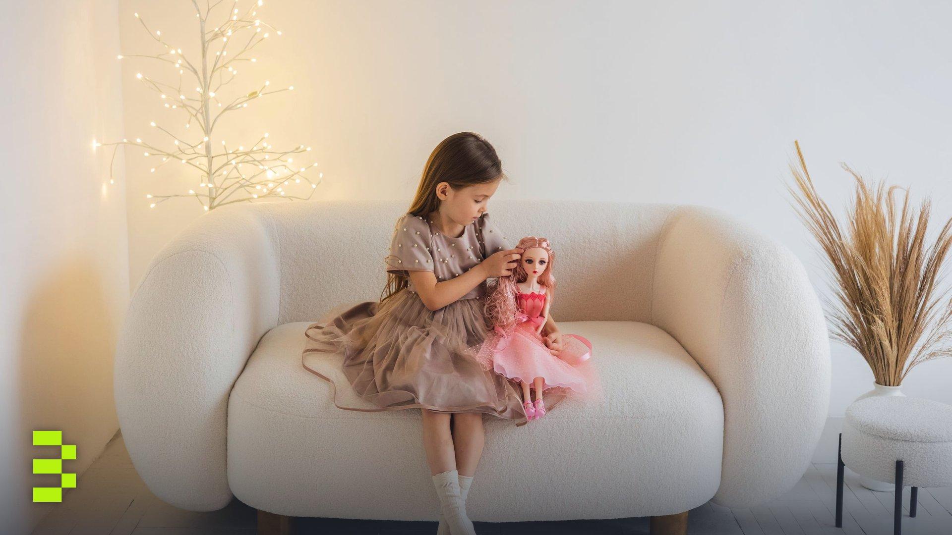 A young girl in a dress sits on a white sofa playing with a pink doll in a minimalist setting.