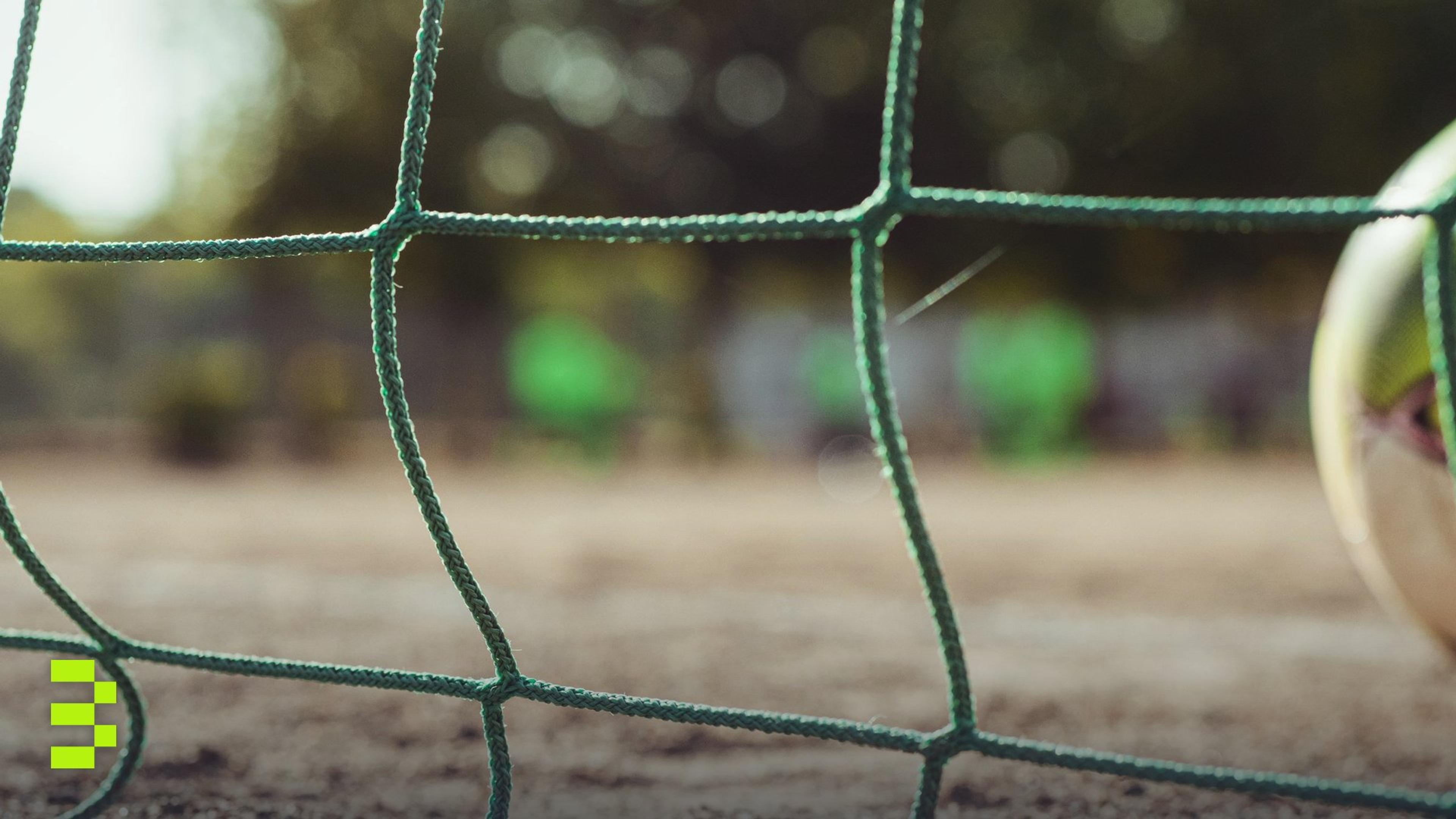Close-up view of a soccer ball near the net on a dry outdoor field with blurred bokeh background.