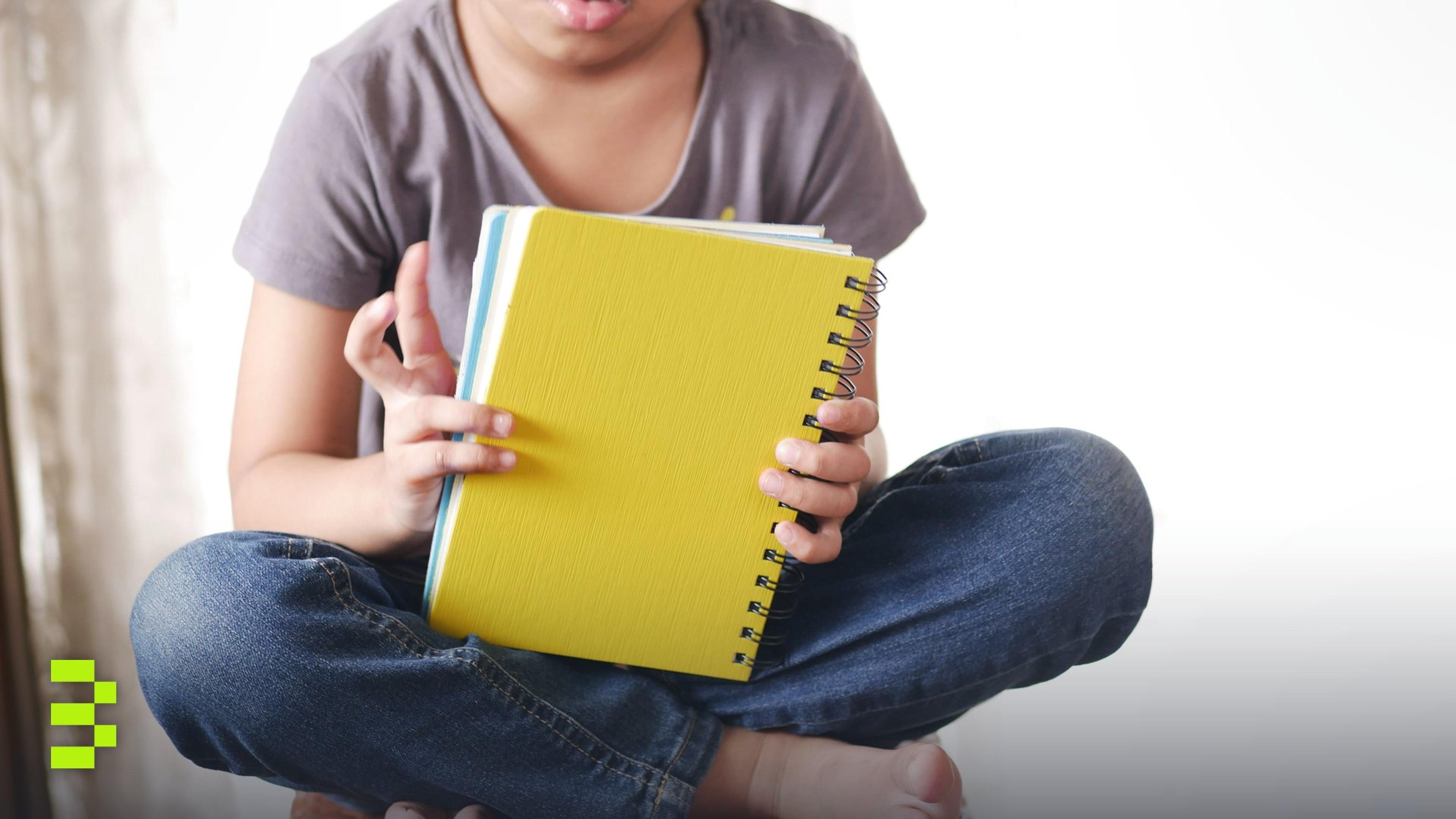 A young girl sitting cross-legged holding a yellow notebook, focusing on reading.