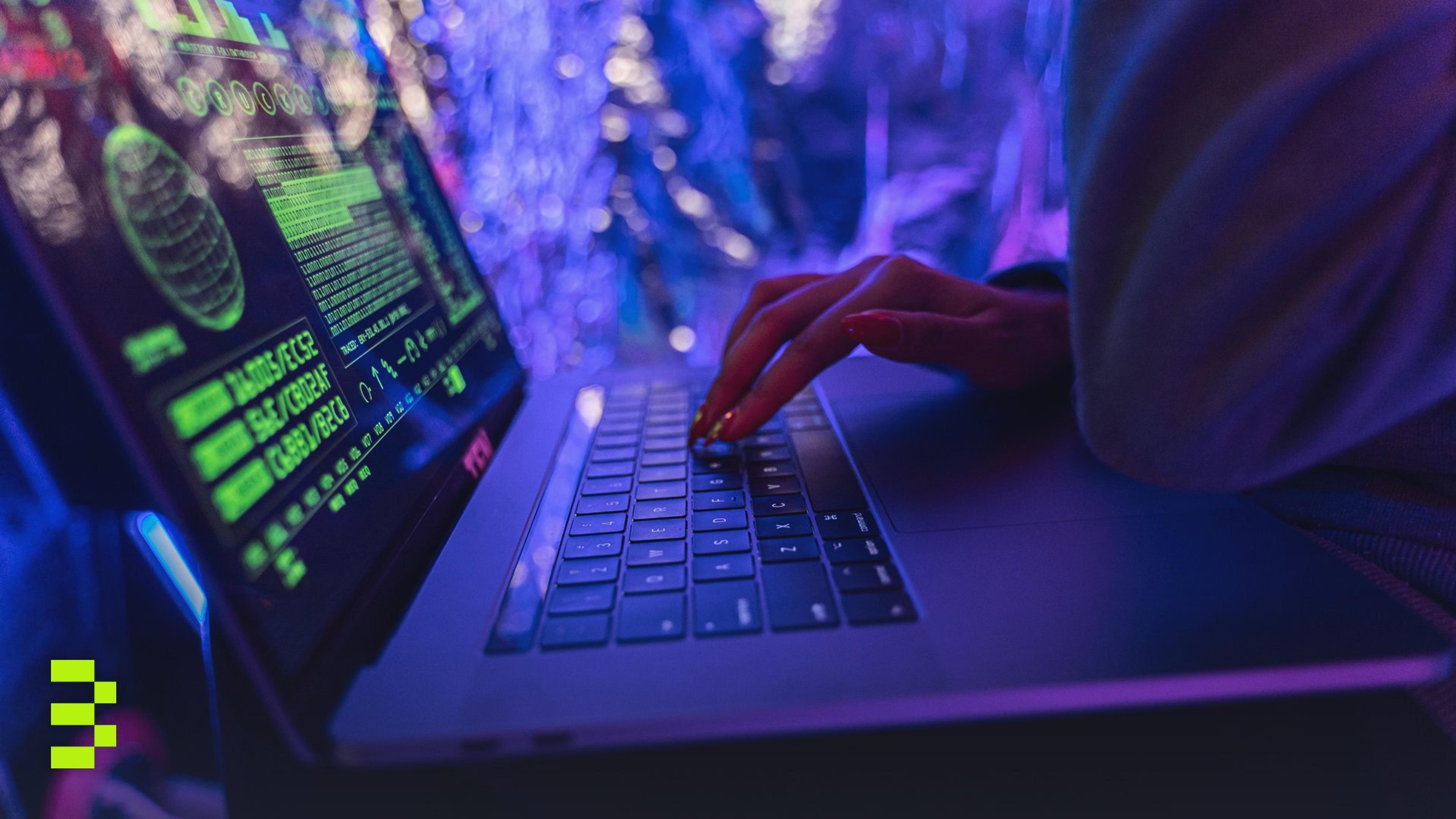 Close-up of hands typing on a laptop displaying cybersecurity graphics, illuminated by purple light.