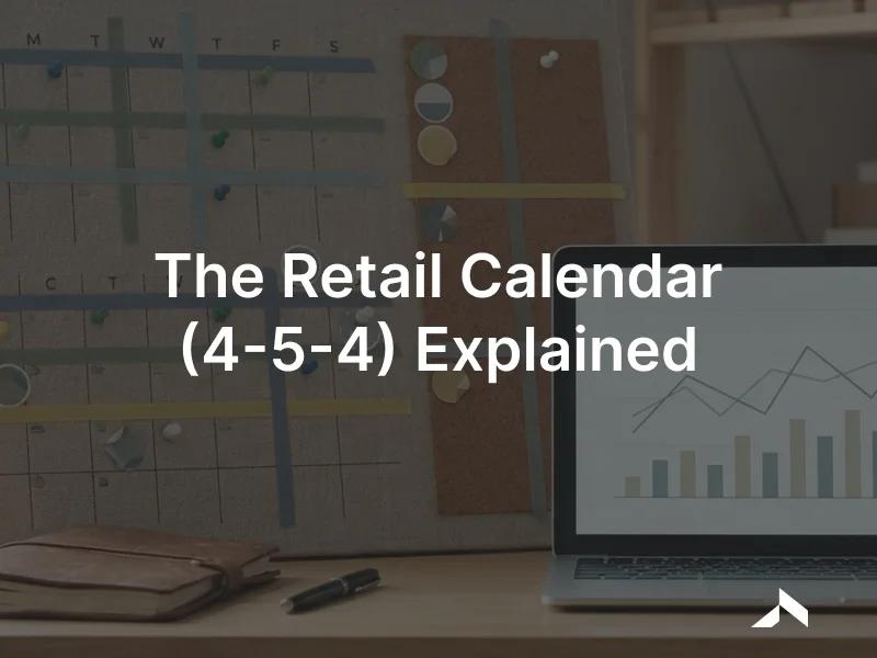 A wall planning calendar of weekly blocks beside a laptop showing a retail sales chart on a desk