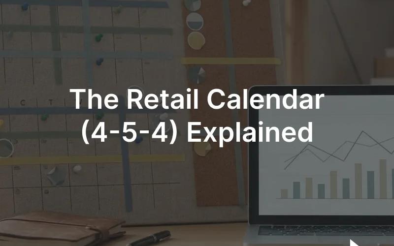 A wall planning calendar of weekly blocks beside a laptop showing a retail sales chart on a desk