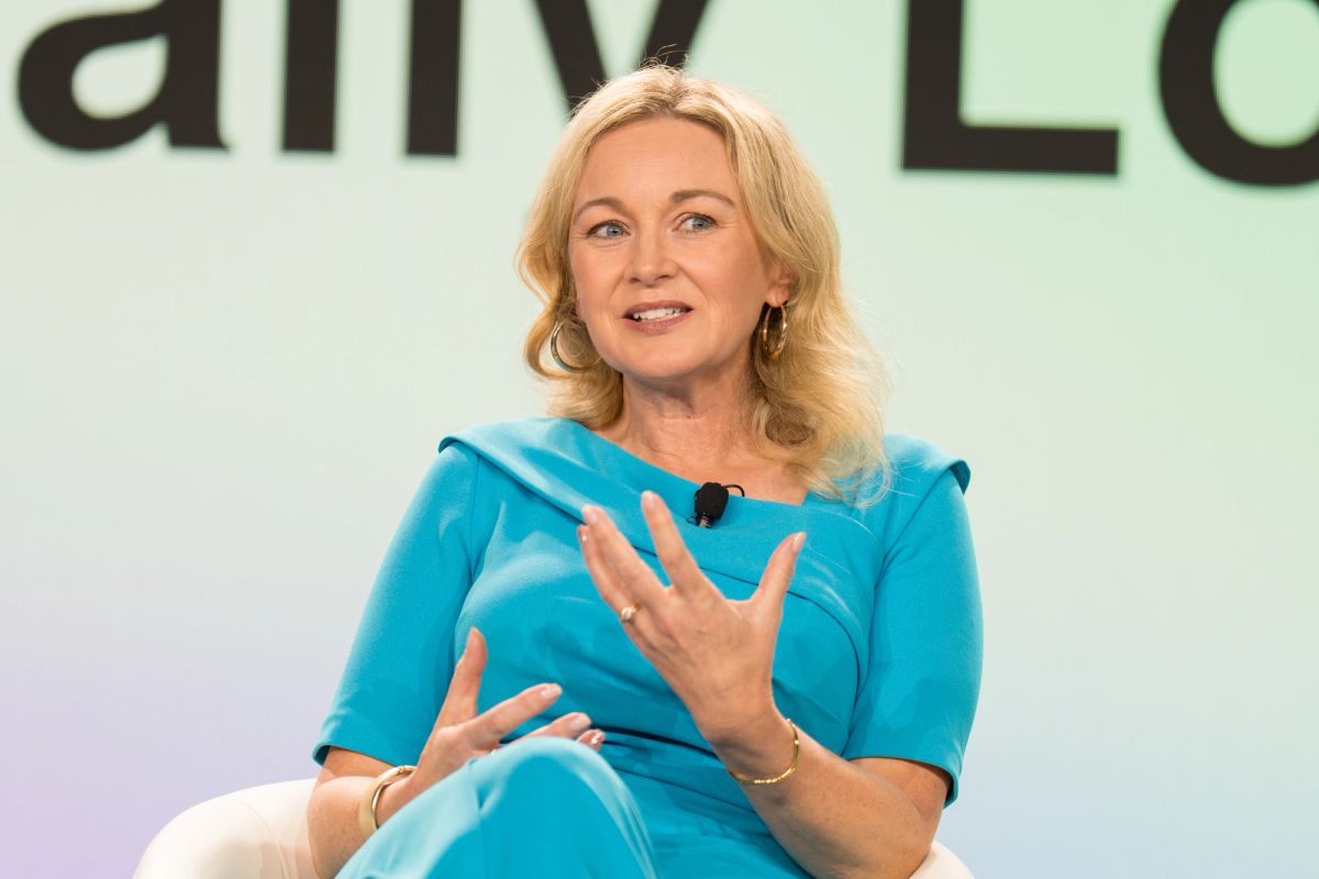 Woman in teal dress speaks at conference panel, gesturing with hands, seated on white chair.