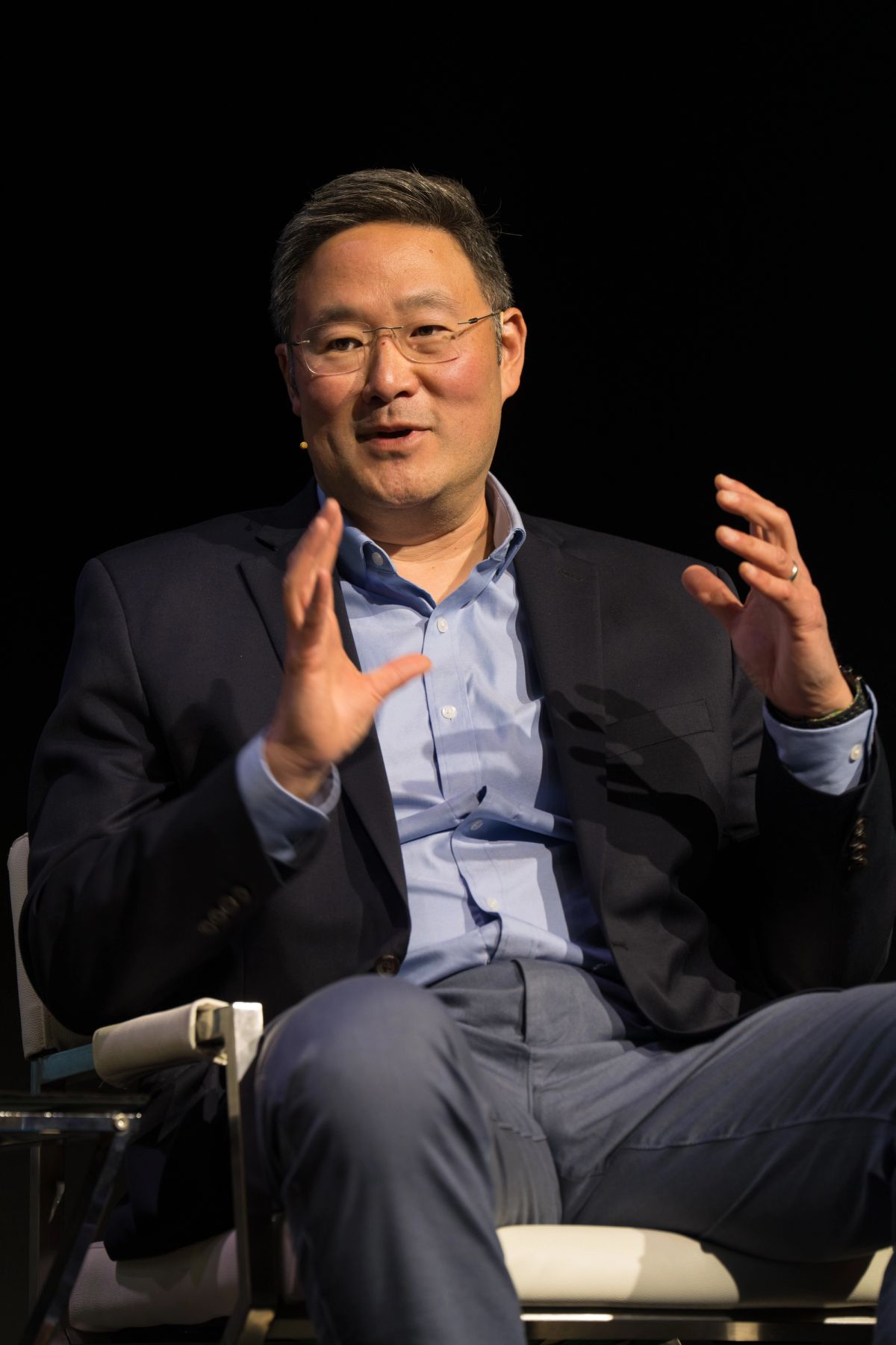 Business executive speaking animatedly with hand gestures while seated on stage at conference event.