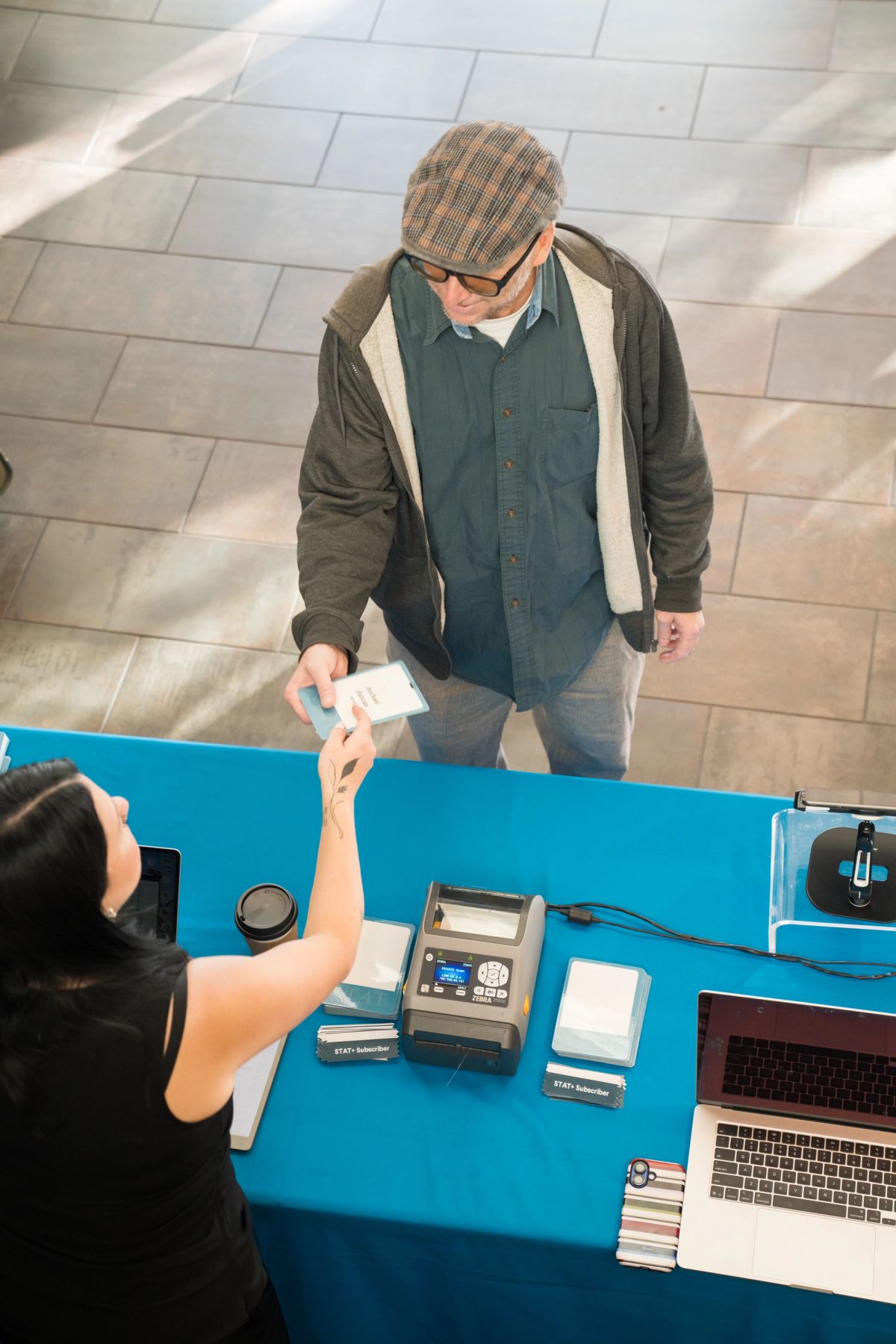 Man in cap and glasses hands document to seated woman at blue table with laptop and electronic devices