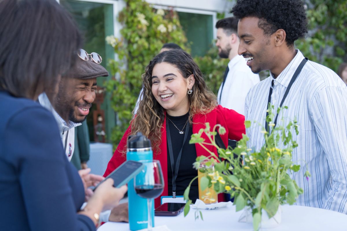 Attendees at an outdoor networking event laugh and chat around a table with plants and refreshments