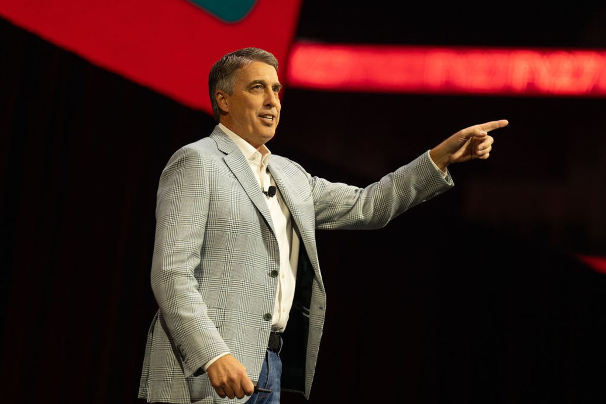 Speaker in gray suit gesturing while presenting on stage with red lighting in background