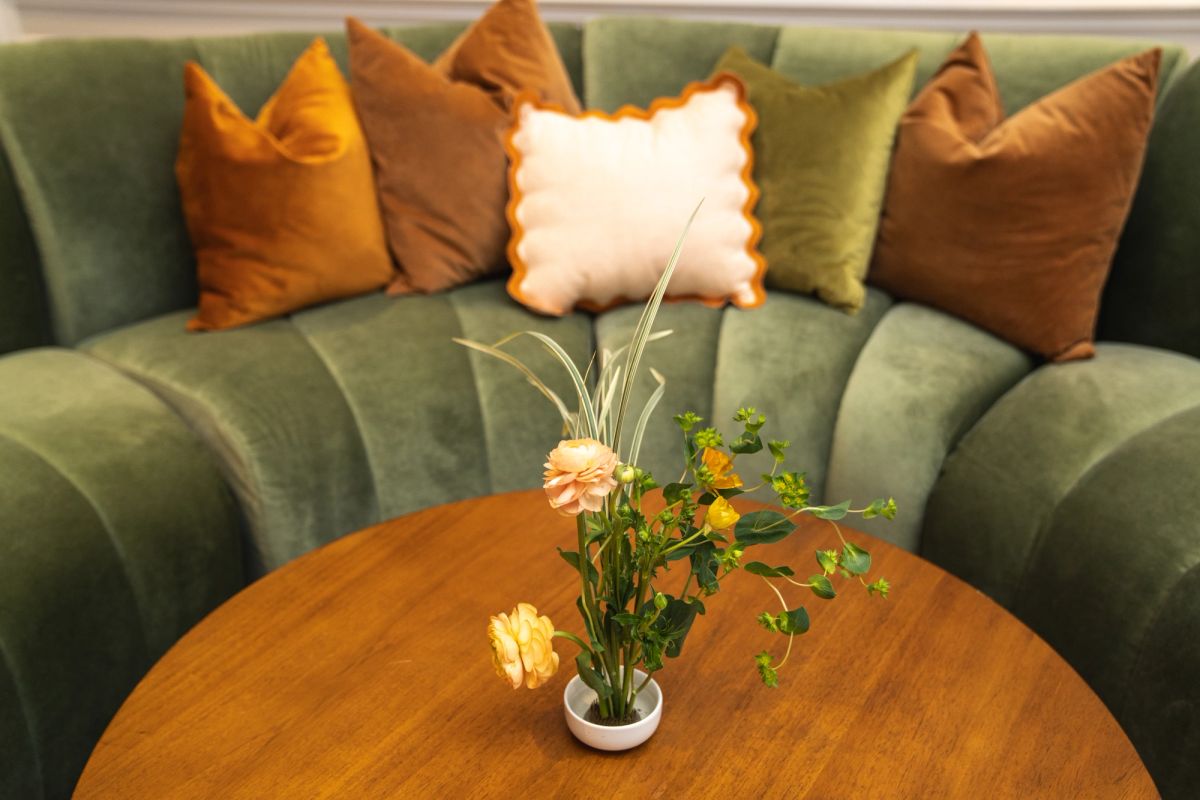 Green curved sofa with orange and white pillows behind round wooden coffee table with small flower arrangement