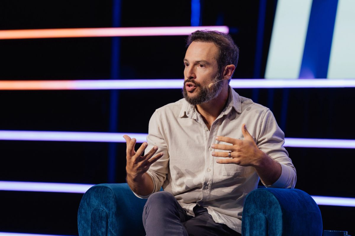 Speaker with beard gesturing while seated in blue chair on stage with colorful lighting at conference
