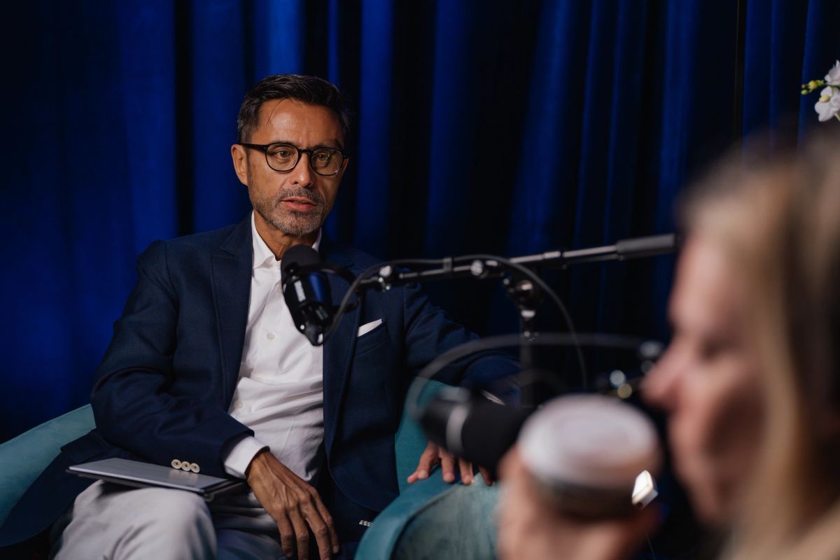 Speaker in glasses and dark jacket sits at microphone during panel discussion with blue curtain backdrop