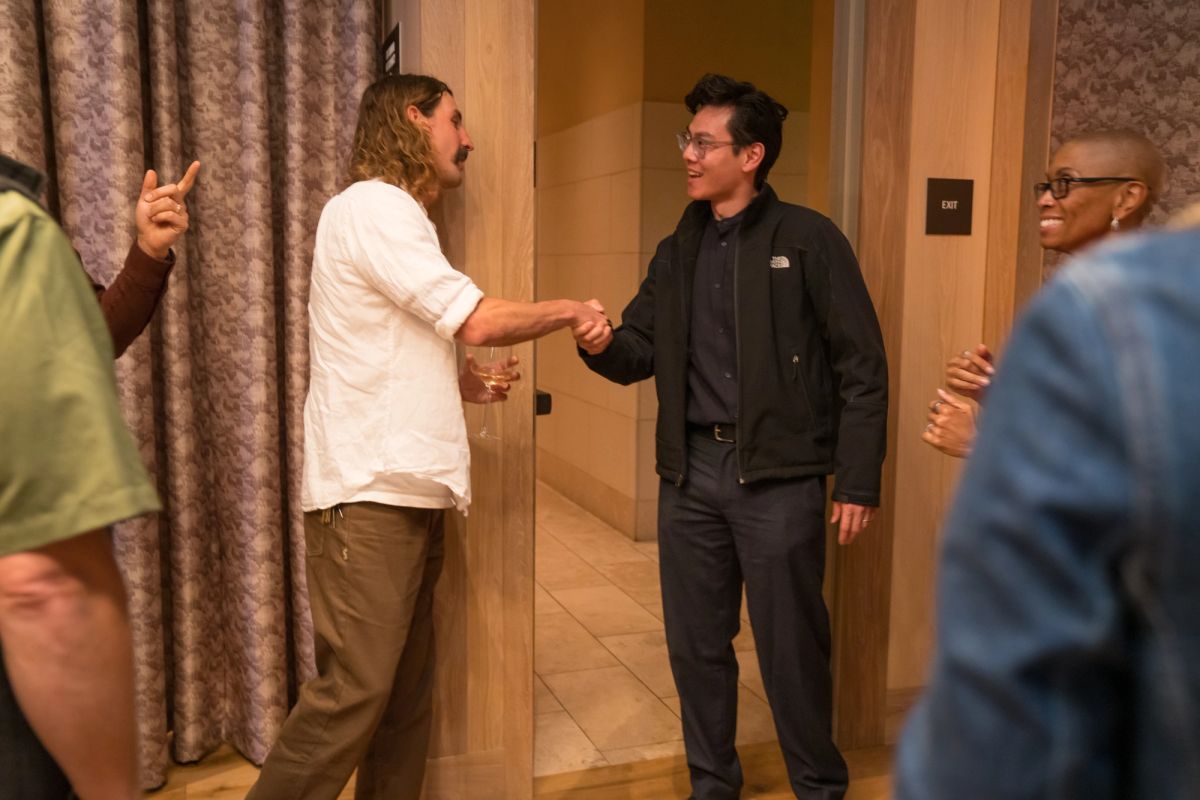 Two men shake hands while others watch in a hotel hallway with patterned curtains and tiled floors.