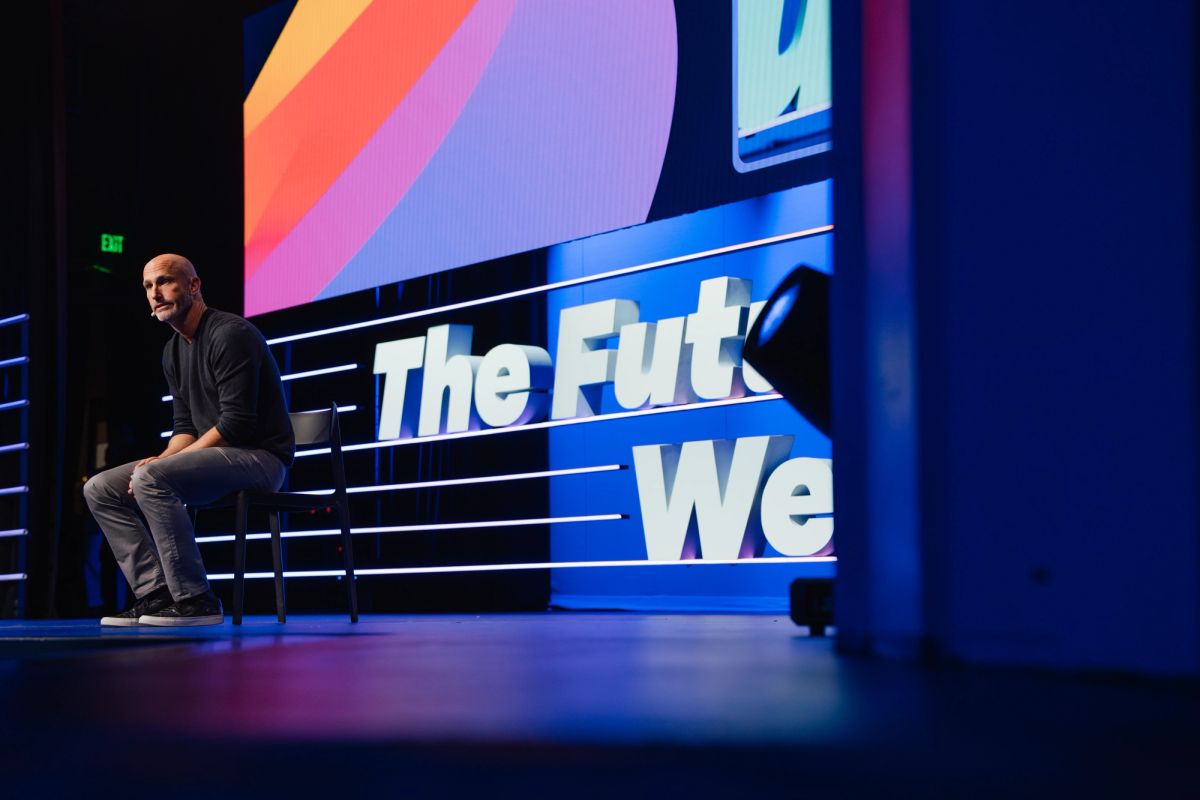 Speaker sits on stage steps beside large blue "The Future We" text display at conference venue