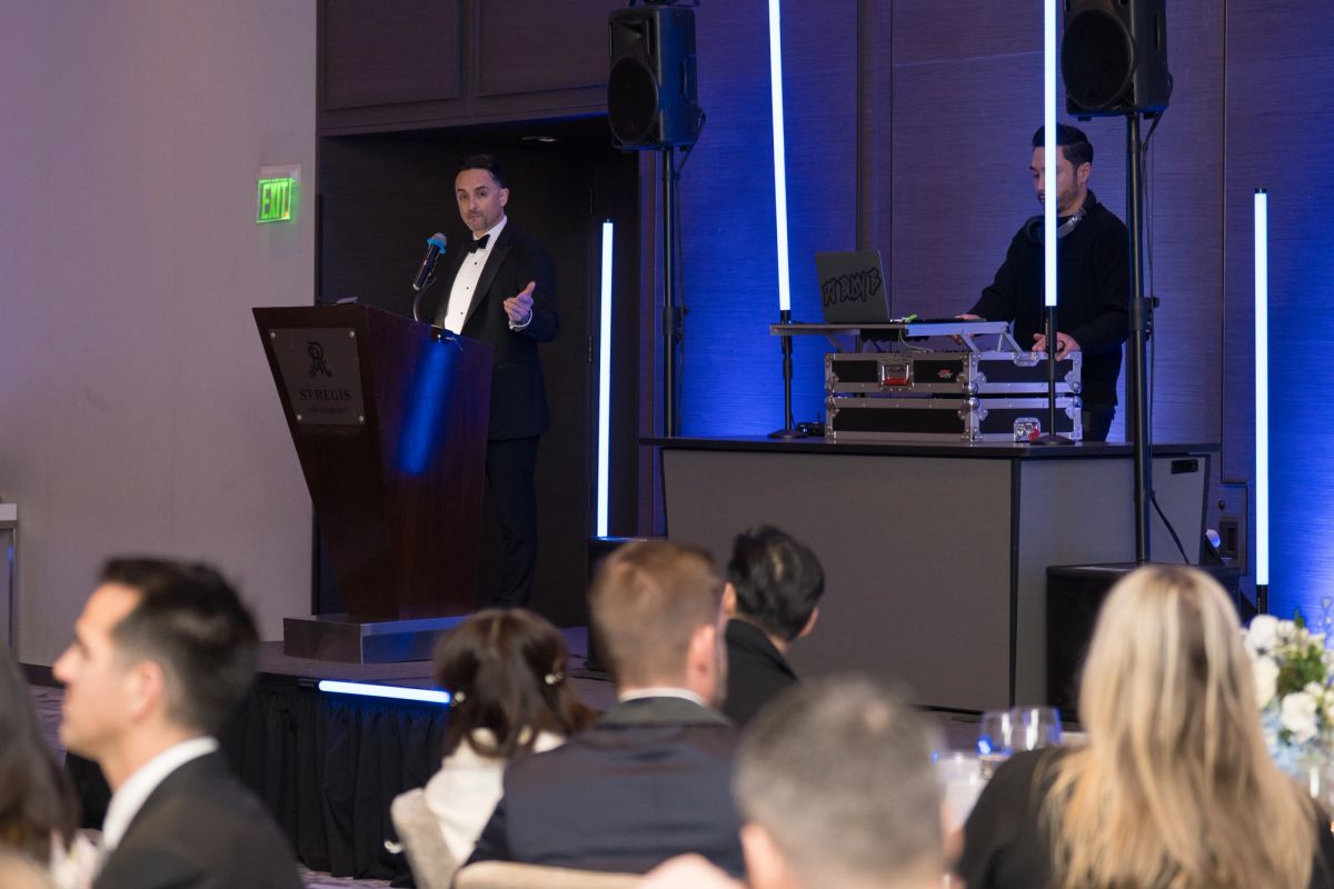 Speaker in tuxedo addresses formal dinner audience with DJ booth visible, blue lighting illuminates event.
