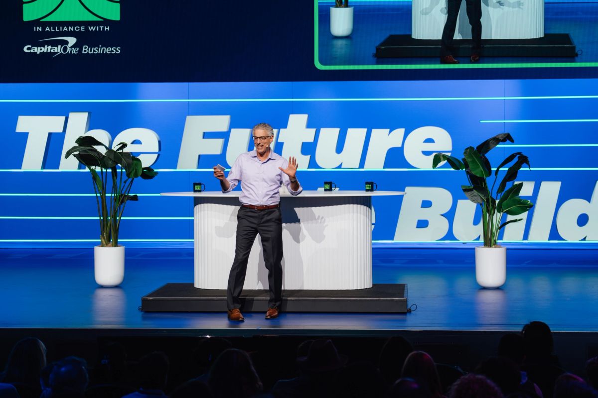 Speaker presenting on stage with "The Future to Build" backdrop, flanked by potted plants at conference venue