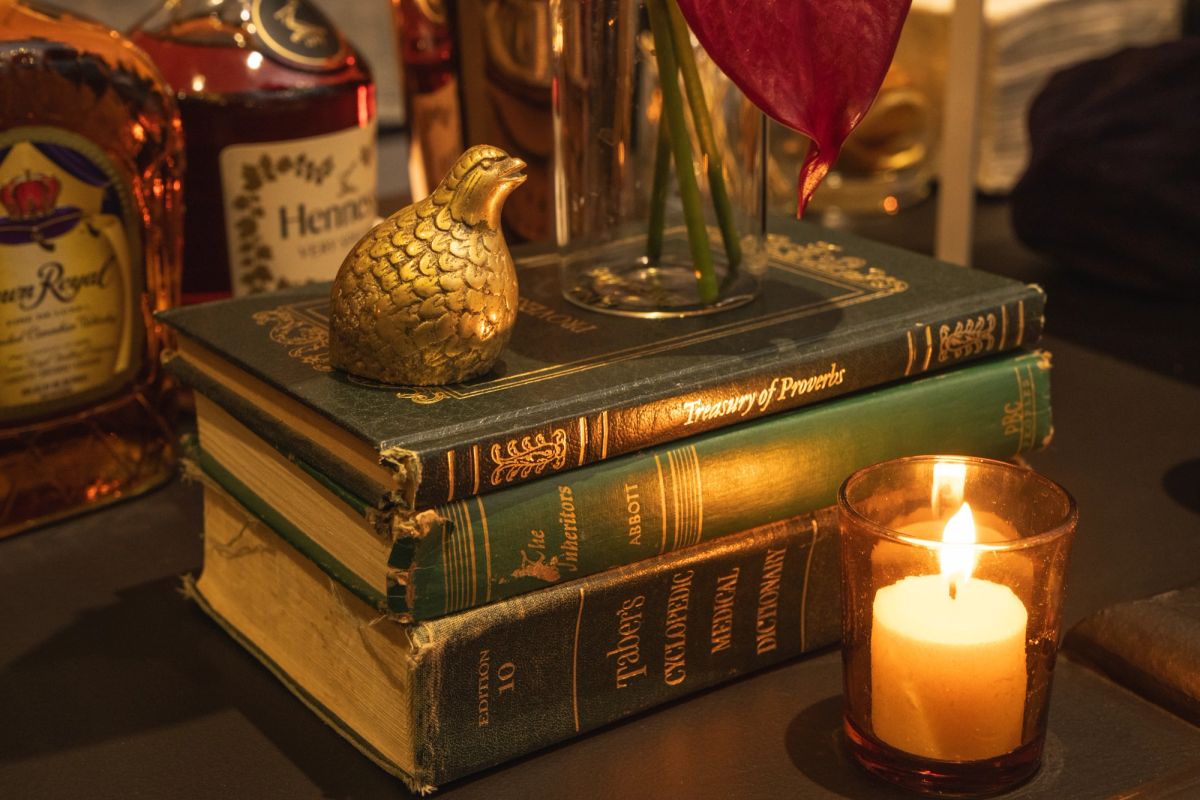 Stack of vintage books with decorative golden bird figurine, lit candle in glass holder, and whiskey bottles.